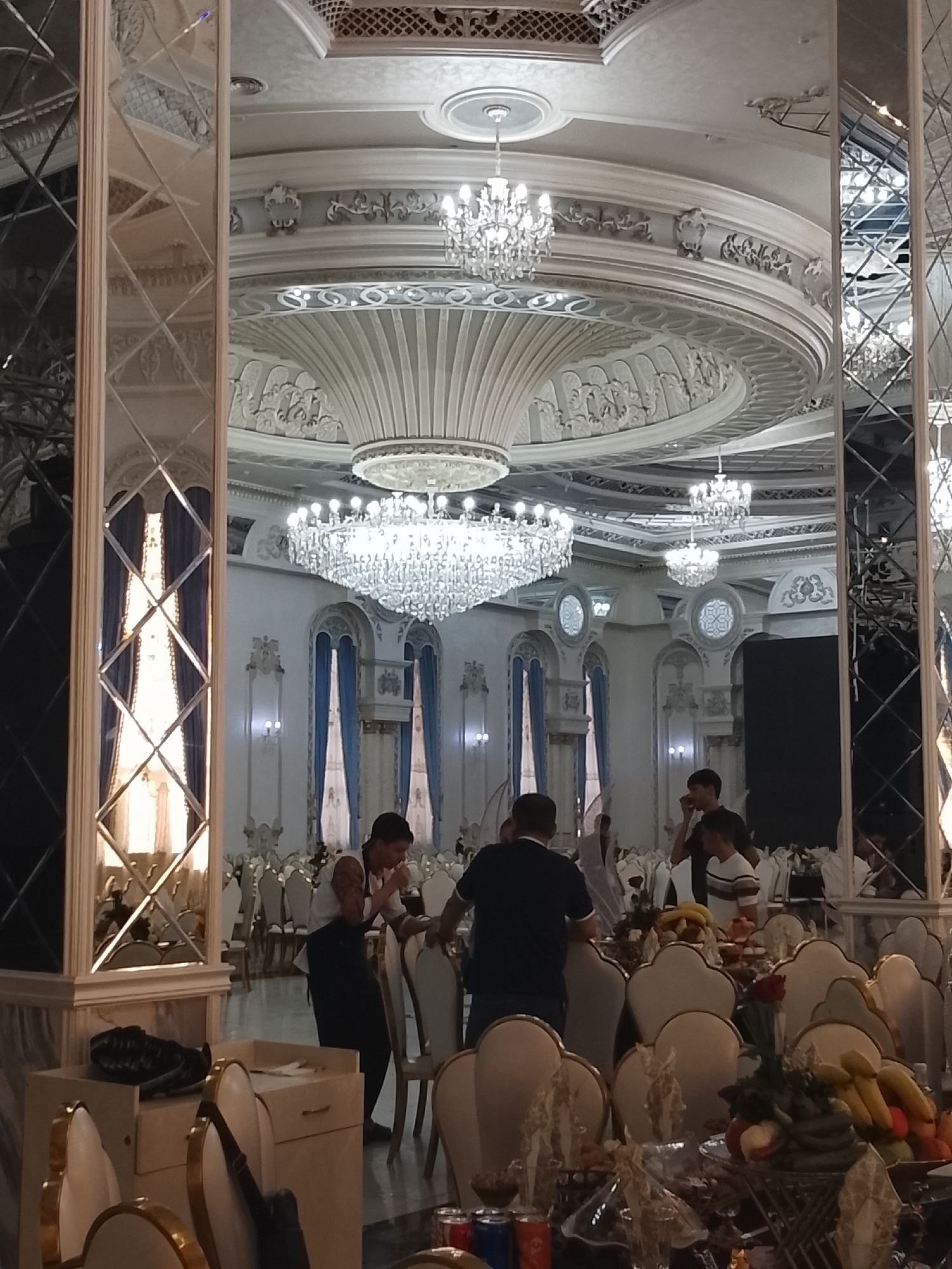 The image depicts the interior of a large, ornate banquet hall, likely in Urgench, Uzbekistan. The hall is elaborately decorated with intricate plasterwork on the ceiling and walls, featuring decorative moldings and a large, central dome. Several crystal chandeliers hang from the ceiling, illuminating the space. Tall, arched windows with blue curtains are visible in the background, contributing to the opulent atmosphere.
In the foreground, a table is set with various food items, including fruits like bananas and apples, as well as vegetables and other decorative elements. Several people are present in the hall, some standing and some seated at tables which are partially obscured by the foreground. One person, a male, is seen wearing a white shirt and dark trousers, appearing to be serving or attending to guests. Another person, a male, is seen wearing a dark t-shirt. The scene suggests a formal gathering or celebration.
The hall also features large, decorative mirrored pillars that reflect the surrounding environment, adding to the sense of space and grandeur. The flooring appears to be dark and polished. The overall impression is one of a luxurious and well-appointed venue for events.