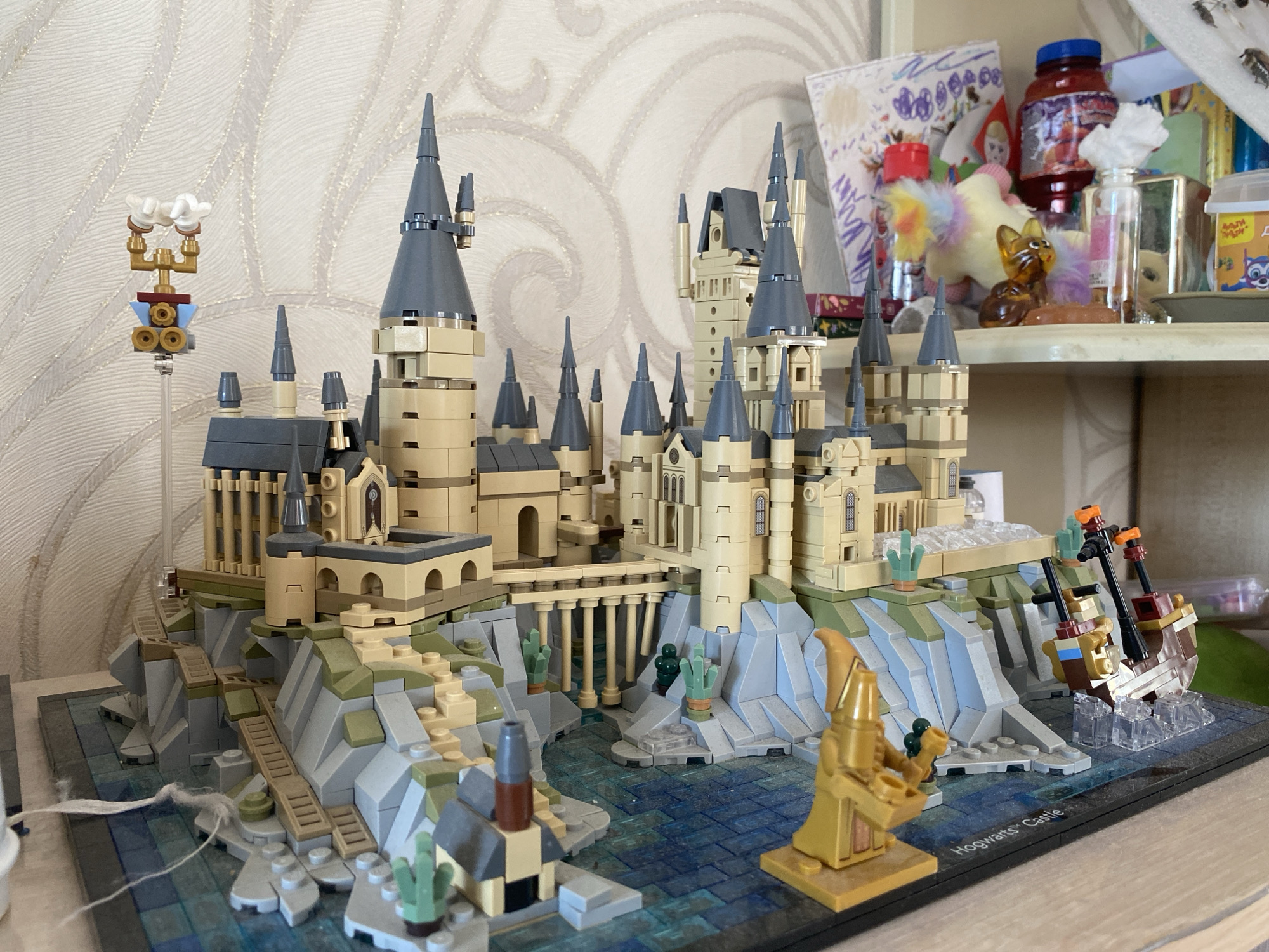 A LEGO model of Hogwarts Castle, likely from the Harry Potter series, is displayed on a dark gray baseplate. The castle is intricate, featuring multiple towers with gray conical roofs, tan brickwork, and arched windows. It is built on a rocky, cliff-like foundation with green and gray LEGO pieces, representing vegetation and stone. A blue and green tiled water area is depicted in front of the castle, with a small rowboat on the right side. A gold-colored LEGO minifigure, resembling a wizard or sorcerer, stands on the right foreground, holding what appears to be a green orb and a golden goblet. There is a smaller building with a chimney on the left foreground. In the background, on a shelf, are various items including a jar with a red lid, a small plush toy with rainbow-colored fur, a glass bottle, and other miscellaneous objects. The wallpaper behind the castle has a textured, light-colored pattern. The scene is indoors, on a table or shelf, and appears to be a personal display. The image does not contain any elements that indicate it is located in the city of Volzhsk, Russia.