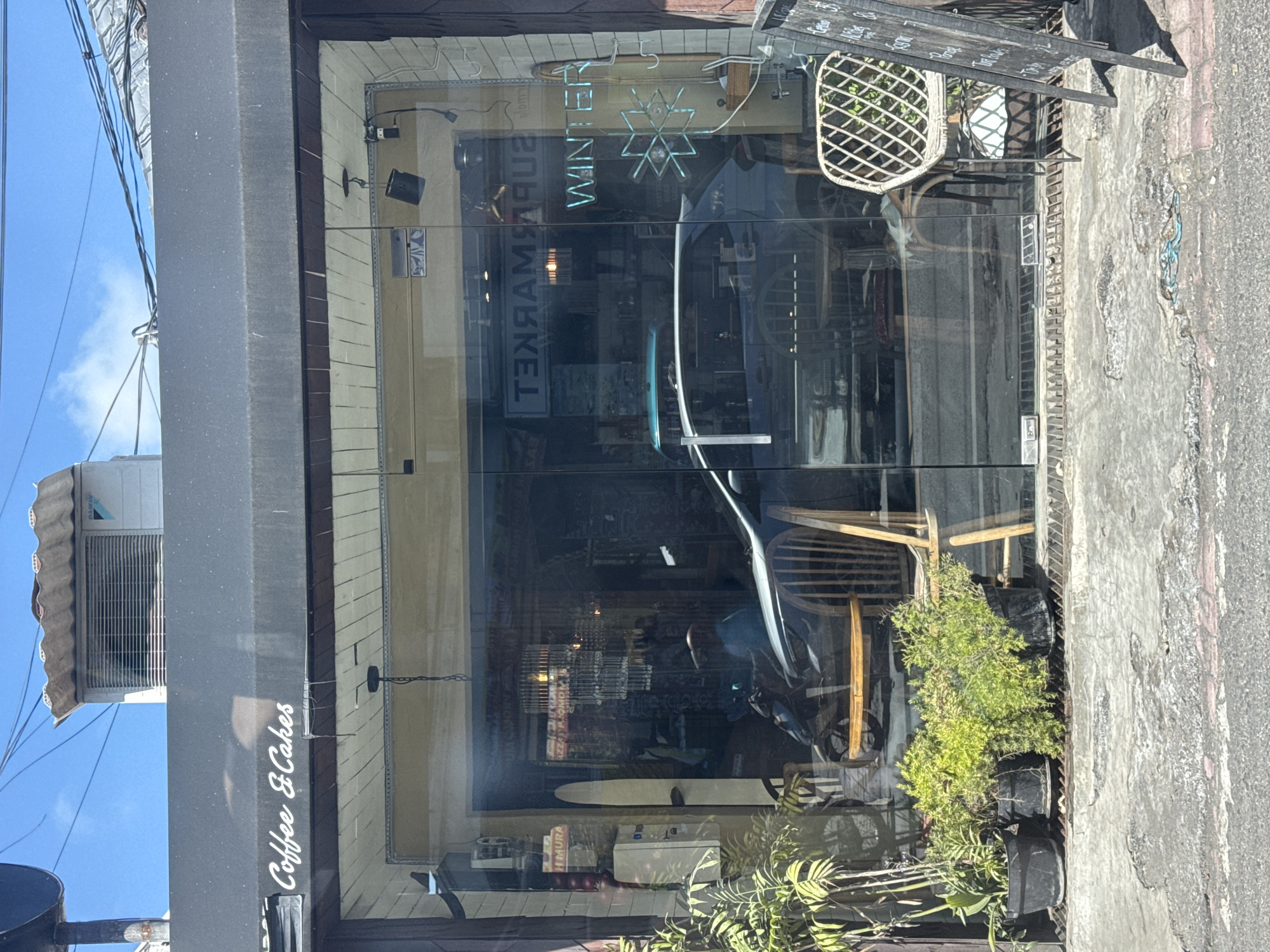 The image shows the storefront of a coffee shop named "Coffee & Cakes" in Kuta Selatan, Indonesia. The shop has a large glass window that reflects the street and some of the interior. Inside, a dark-colored car is parked, and a person is visible on a motorcycle. A neon sign that reads "WINTER" is illuminated in the window, along with a snowflake graphic. Plants in pots are placed in front of the shop. The exterior of the building is visible above the awning, with an air conditioning unit mounted on the wall. The street in front of the shop is paved, with some cracked areas. A chalkboard sign stands to the right of the entrance. The sky above is blue with scattered clouds.