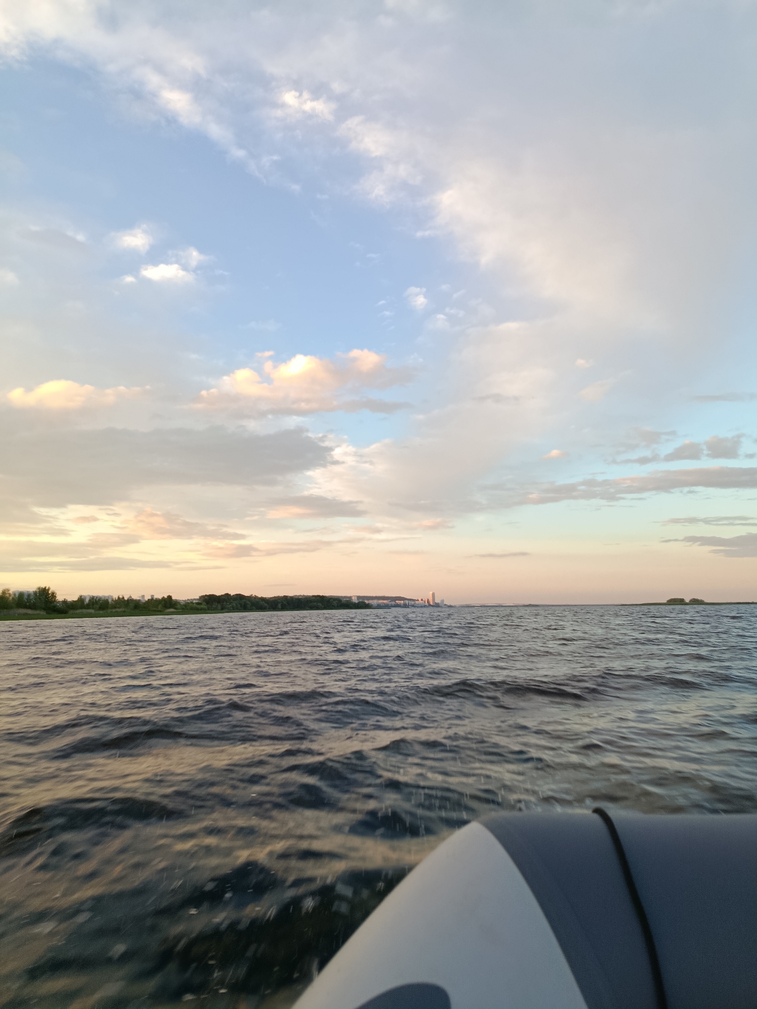 A wide-angle shot captures a scenic view of the Volga River in Saratov, Russia, as seen from a boat. The foreground is dominated by the dark, rippling surface of the river, with a portion of a grey and white inflatable boat visible in the lower right corner. In the distance, a lush green shoreline stretches across the horizon, dotted with trees and punctuated by several buildings, suggesting the presence of a city or town. Above, a vast sky displays a dynamic arrangement of clouds, transitioning from a pale blue at the top to warmer, pastel tones of pink and orange near the horizon, indicating either sunrise or sunset. The overall scene evokes a sense of calm and natural beauty.