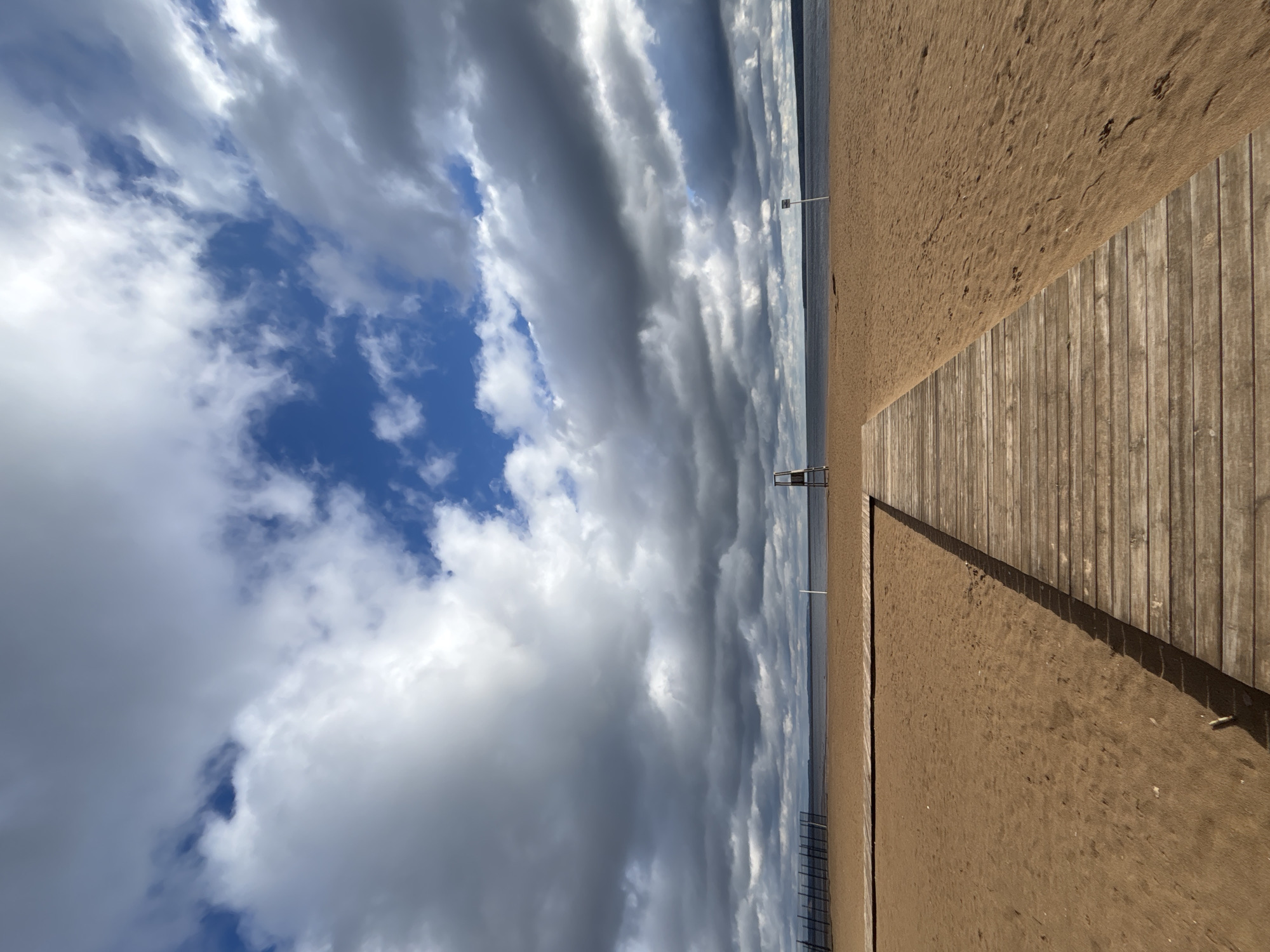 The image captures a peaceful, unpopulated beach scene in Chaikovsky, Russia, under a dramatic sky. The upper portion of the frame is dominated by a vast sky filled with elongated, striking clouds, ranging from bright white to dark grey, interspersed with patches of clear blue. This cloud formation creates a sense of depth and movement.
On the right side, a light-colored wooden walkway or deck, composed of vertical planks, stretches into the distance, leading the eye towards the horizon. This walkway casts a long, distinct shadow onto the adjacent sandy beach, suggesting the sun is positioned to the side, likely indicating late morning or afternoon. The sandy beach itself appears pristine and undisturbed, devoid of footprints or any human activity.
The sand meets a calm, expansive body of water on the horizon, which appears as a flat, blue-grey line under the cloud cover. In the distance, on the sandy shore, a white pole is visible, along with what appears to be a lifeguard chair or similar elevated structure further down. Faint lines suggesting a fence or posts can also be seen closer to the water's edge in the mid-ground. The overall scene conveys a feeling of tranquility and the raw beauty of nature, with no visible text.