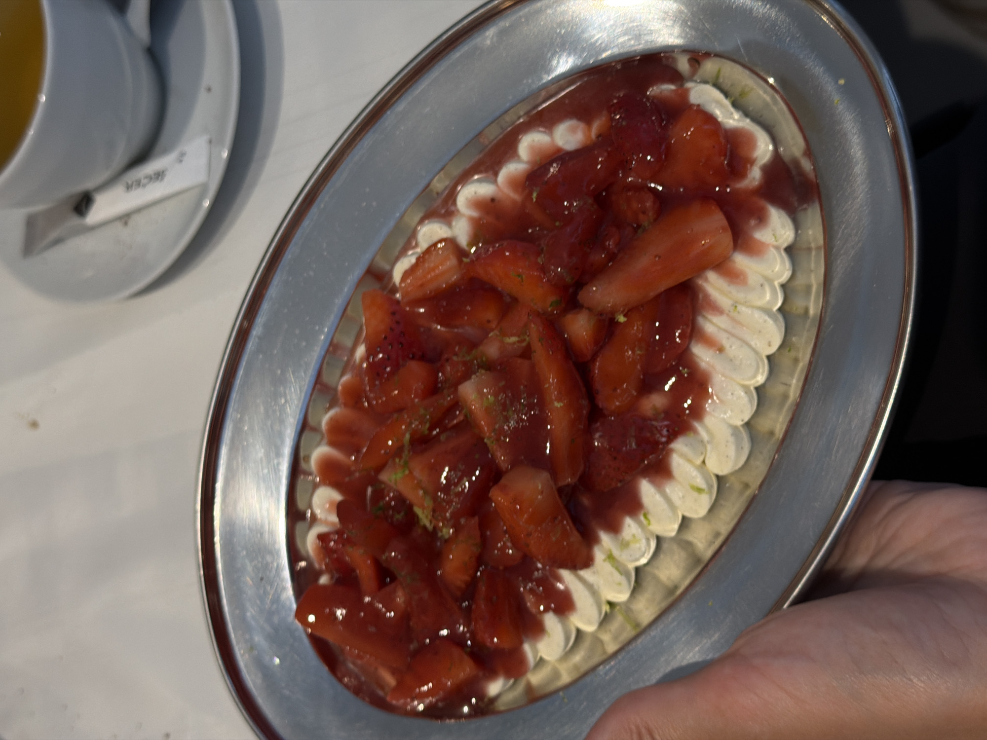 A close-up shot reveals a silver oval platter holding a dessert of strawberries and cream, positioned alongside a white cup and saucer, with a sachet of sugar bearing the word "Šećer". The strawberries, coated in a glossy red sauce, are arranged atop piped white cream. A person's hand is partially visible, holding the edge of the platter. The setting suggests a café or restaurant setting, likely in Belgrade, Serbia, given the sugar sachet text.