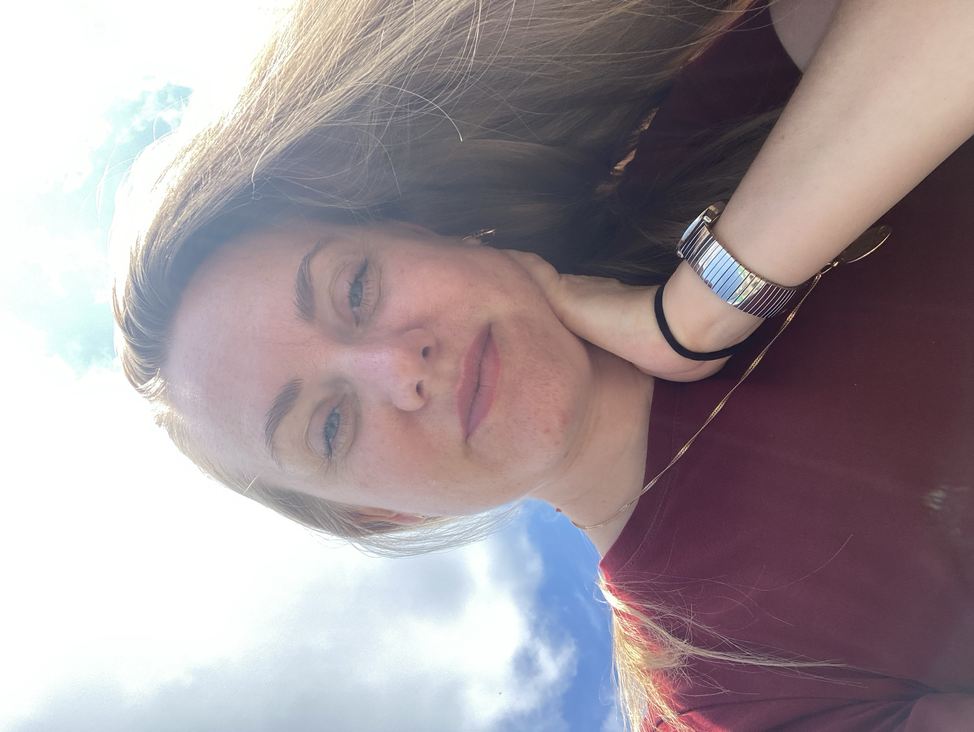 A young woman with long blonde hair is taking a selfie outdoors against a bright, cloudy sky. She is wearing a maroon t-shirt and a gold necklace. Her right hand is resting on her chin, and she is wearing a silver watch on her left wrist. The lighting suggests it is daytime, possibly sunny with some cloud cover. There is no discernible location within the city of Марковский, Russia.