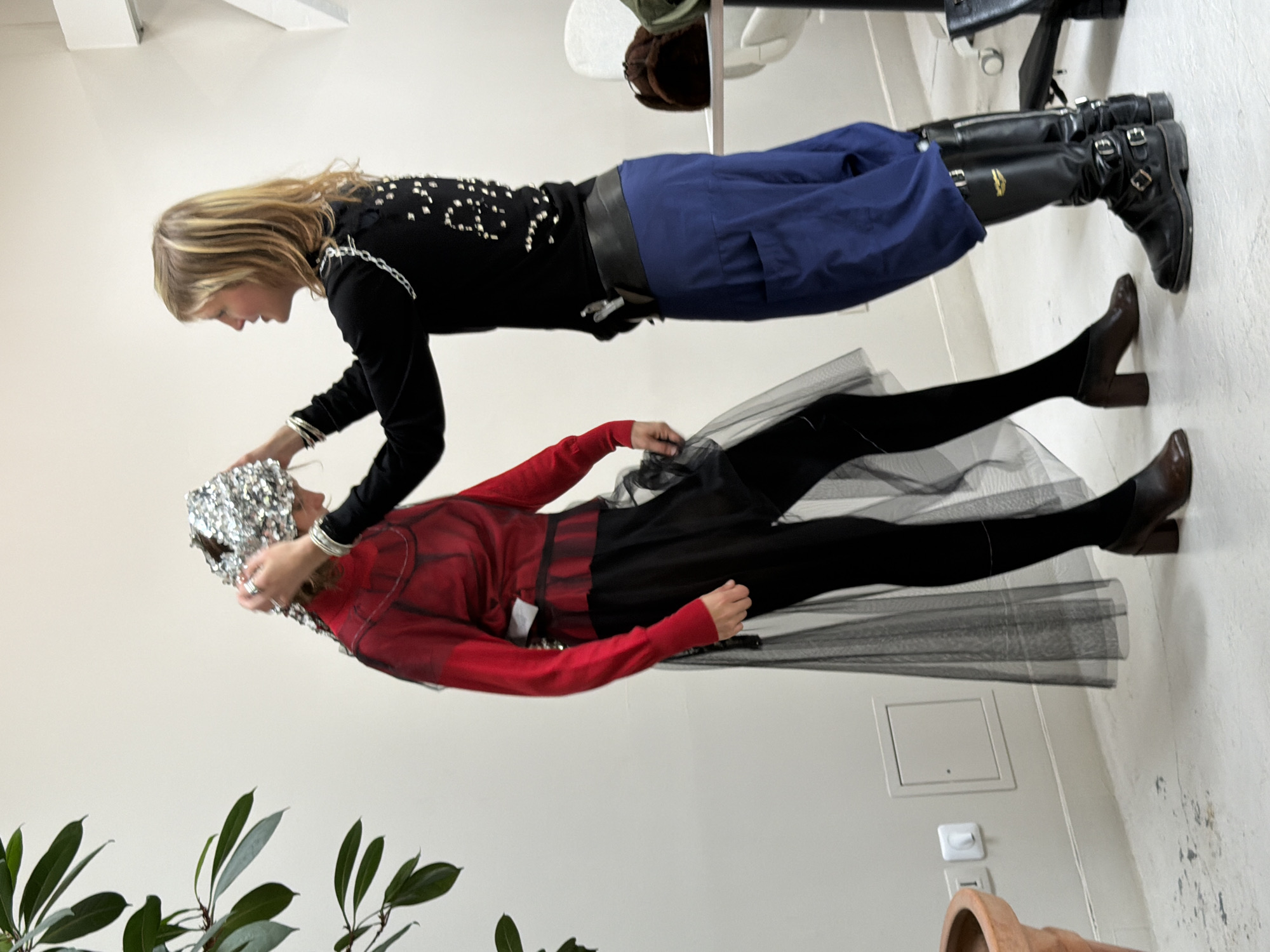 Two individuals are positioned indoors against a stark white wall. One is attired in a black, long-sleeved top adorned with white decorative elements, coupled with a wide black belt, blue trousers, and black boots. They are leaning forward, their hands appearing to adjust a silver, foil-like head covering on the second individual.
The second individual is dressed in a red, long-sleeved top, a black skirt, and black leggings, completed with brown heeled shoes. The foil head covering obscures much of their head.
Below, to the left, is a green plant in a terracotta pot. On the right side of the scene, there's an electrical outlet and a utility access panel on the wall. The location is undetermined, but the style of electrical fixture does not suggest Paris, France.