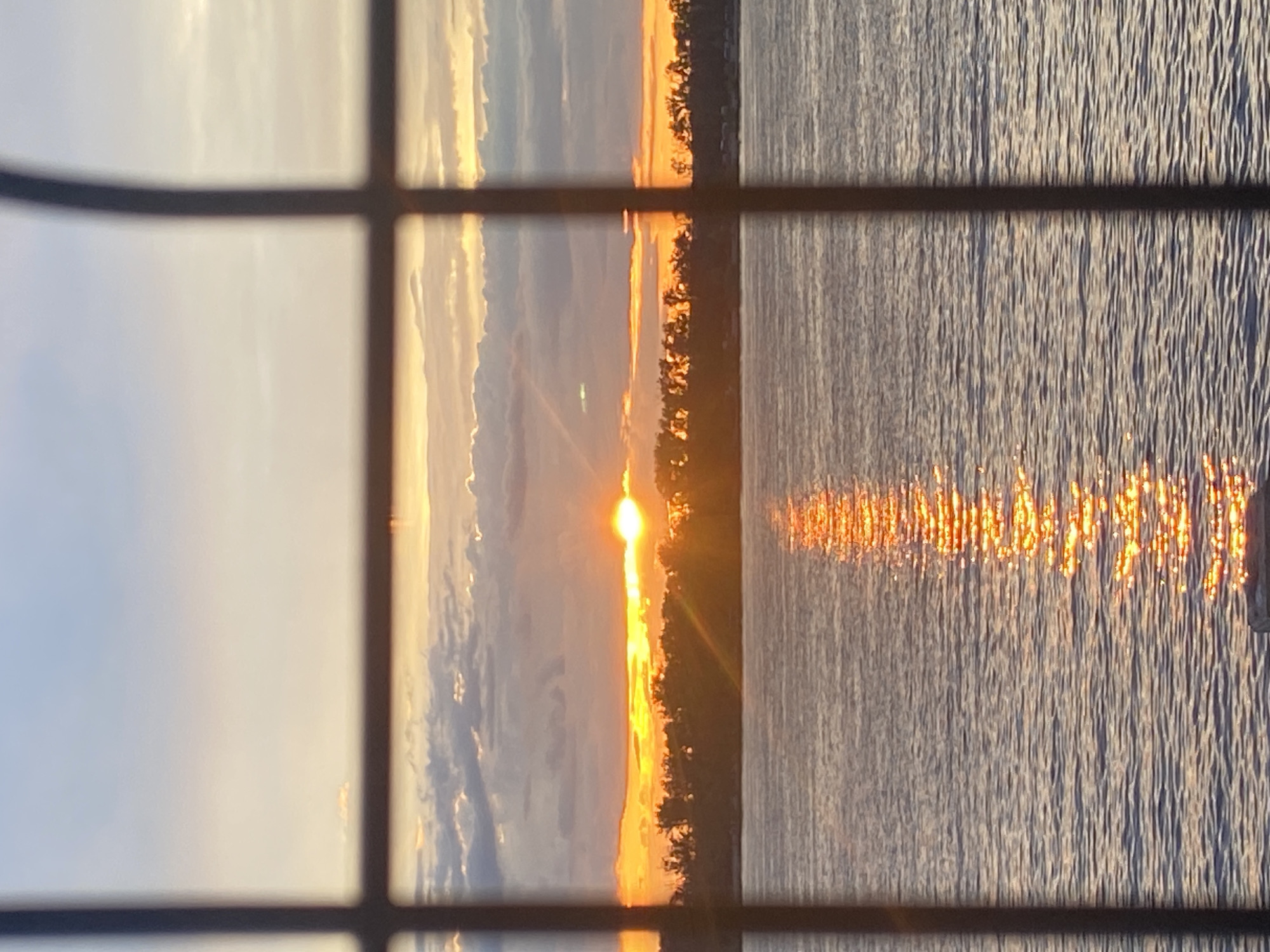 This image depicts a sunset over a body of water, viewed through a dark, grid-like structure, possibly a window or fence. The sky is filled with a gradient of colors, from soft blues and oranges near the horizon to lighter blues at the top, with scattered clouds. The sun is low on the horizon, casting a bright, golden glow and reflecting intensely on the rippling surface of the water. A silhouetted treeline runs along the distant shore. The scene is framed by dark, vertical and horizontal bars, creating a sense of looking out from an enclosed space. There are no people or specific objects in the foreground that are clearly identifiable. The overall atmosphere is serene and picturesque, characteristic of a sunset over a calm body of water. The location is identified as Novokuybyshevsk, Russia.