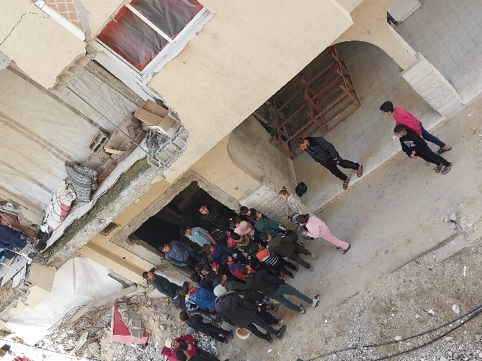 An overhead view depicts a damaged urban street or courtyard in Khan Yunis, Palestinian Territory. Approximately 20-25 individuals, predominantly children, are present amidst the debris of partially collapsed buildings. A large cluster of children, dressed in various winter clothing including jackets and hoodies, stands around a dark, recessed opening in a severely damaged building wall on the left side of the frame. Exposed rebar, concrete rubble, and broken masonry are evident on and around this structure, which shows multiple layers of collapse and internal exposure.
To the right, a multi-story building, also bearing signs of damage with exposed internal walls and a window covered by red material, stands partially intact. On a damaged ledge of this building, items resembling mattresses, blankets, and concrete blocks are visible. The ground is a mix of concrete and scattered rubble, with dark cables running across the lower left foreground.
Further down the path, away from the main group of children, three additional individuals are visible. One adult male, wearing a dark jacket and sandals, stands looking towards the cluster of children. Two younger individuals or teenagers, one in a pink top embracing another in a dark top, stand near a collapsed wooden structure further along the path. A child in a pink tracksuit is seen walking away from the larger group towards these individuals. The scene conveys human presence within a heavily damaged environment.