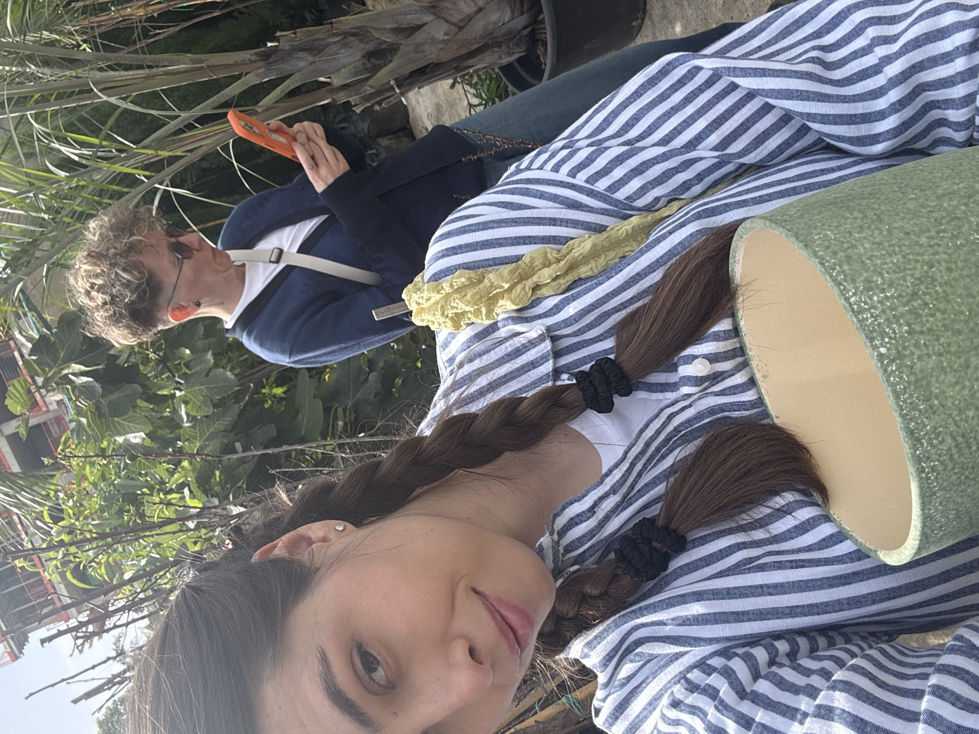 A medium-shot portrait shows a woman wearing a blue and white striped shirt with dark hair in braids held by black hair ties. She holds a light green planter. In the background, partially obscured by tall grasses, is another person with short, curly hair wearing sunglasses, a dark blue jacket, and a white t-shirt. This person holds an orange device. The scene is set outdoors, possibly in a garden or nursery, with buildings and green foliage visible. The location is Tel-Aviv, Israel, as suggested by the urban landscape and vegetation.