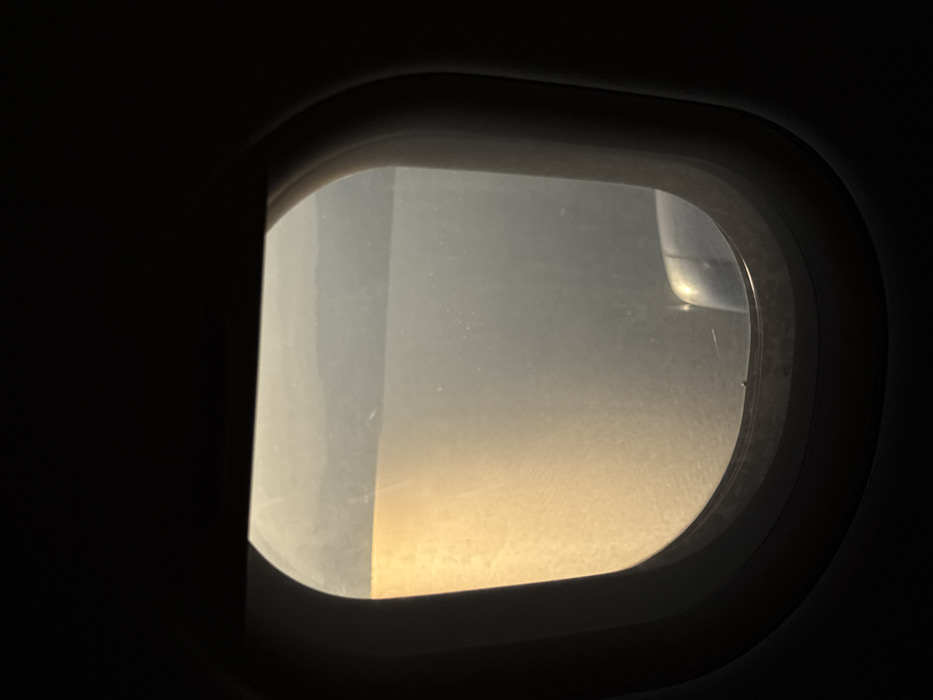 An airplane window view with a blurred landscape outside. Golden light dominates the lower portion of the window, transitioning to a hazy, brighter area above. Part of the airplane wing is faintly visible in the upper right corner. The window frame is dark and prominent, sharply contrasting with the outside light. The interior of the plane surrounding the window is dark. The window glass has visible streaks and imperfections.