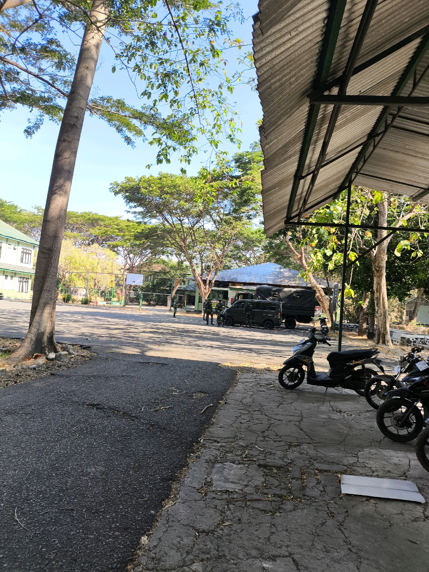 A group of individuals in military uniforms are standing near several military vehicles, including a truck and a van, in what appears to be an outdoor area with buildings and trees. The ground is paved with asphalt and concrete. Several motorcycles are parked on the concrete area in the foreground, under a corrugated metal roof structure. The scene is illuminated by sunlight, creating shadows on the ground. The environment suggests a military base or training facility in Unknown, Indonesia.