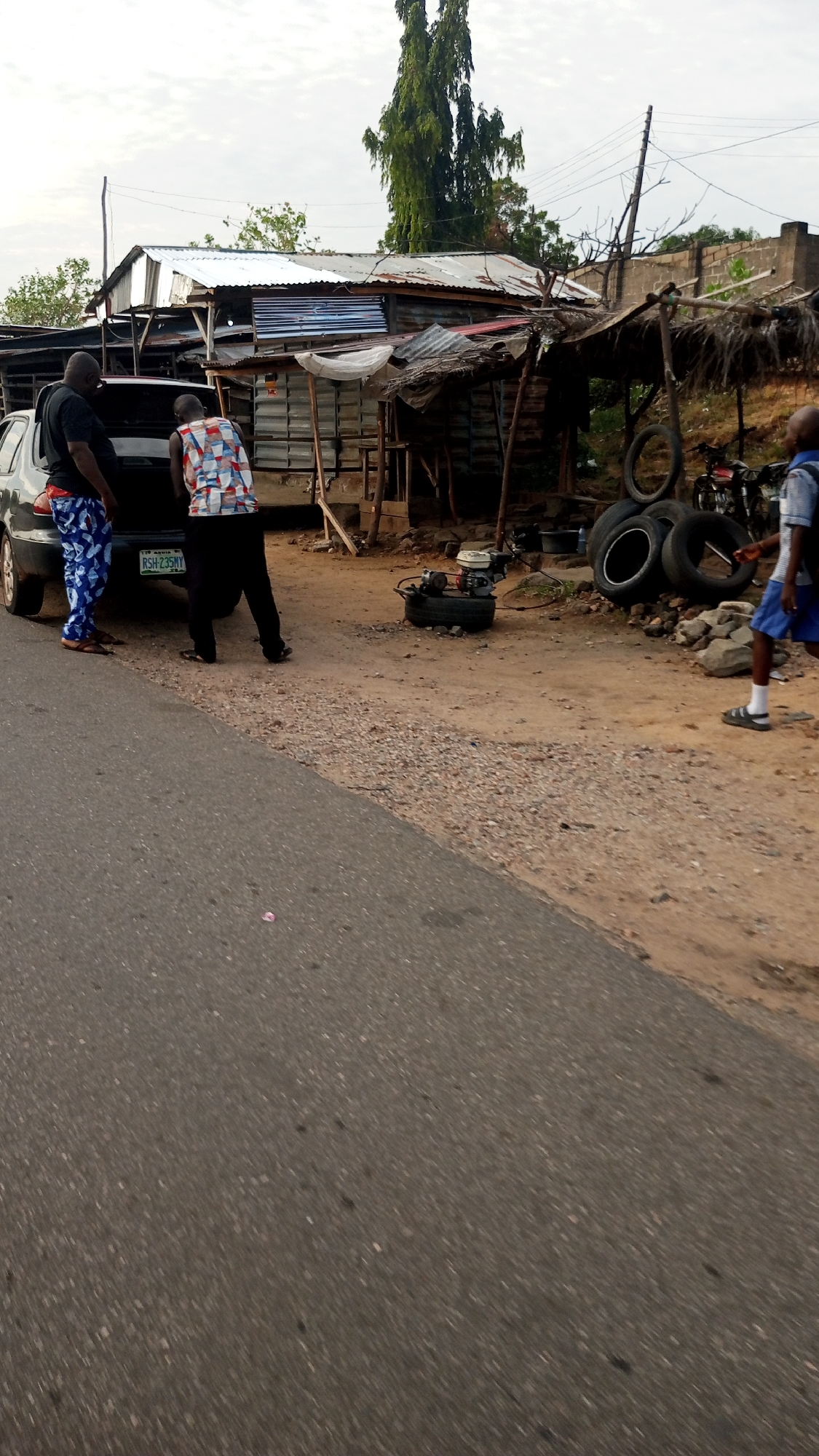The image presents a street scene in Lokoja, Nigeria, under an overcast sky. In the foreground, a paved road angles diagonally across the lower left. Adjacent to the road is a dirt and gravel shoulder.
Two men are standing at the rear of a dark-colored car, which is parked on the dirt shoulder. The car's trunk is open, and both individuals are looking inside, possibly in the process of loading or unloading items. The man on the left wears a dark t-shirt and blue patterned trousers, while the man on the right is dressed in a multi-colored patterned shirt and dark trousers. The car's license plate, "RSH-235MY," is clearly visible, with "179 Abuja W" printed above it, indicating Nigerian registration.
Behind the car, several rustic structures with corrugated iron roofs are visible, suggesting roadside shops or workshops. An engine or generator unit rests on a car tire on the ground to the right of the car. Further right, a stack of old tires and a small shed with a rough roof are present, with two motorcycles partially visible behind them. A young boy, wearing a blue and white striped shirt, blue shorts, white socks, and sandals, is walking towards the left side of the frame in the mid-ground.
A tall, slender tree dominates the background against the cloudy sky, and power lines are visible overhead. The scene depicts an active, informal roadside environment.