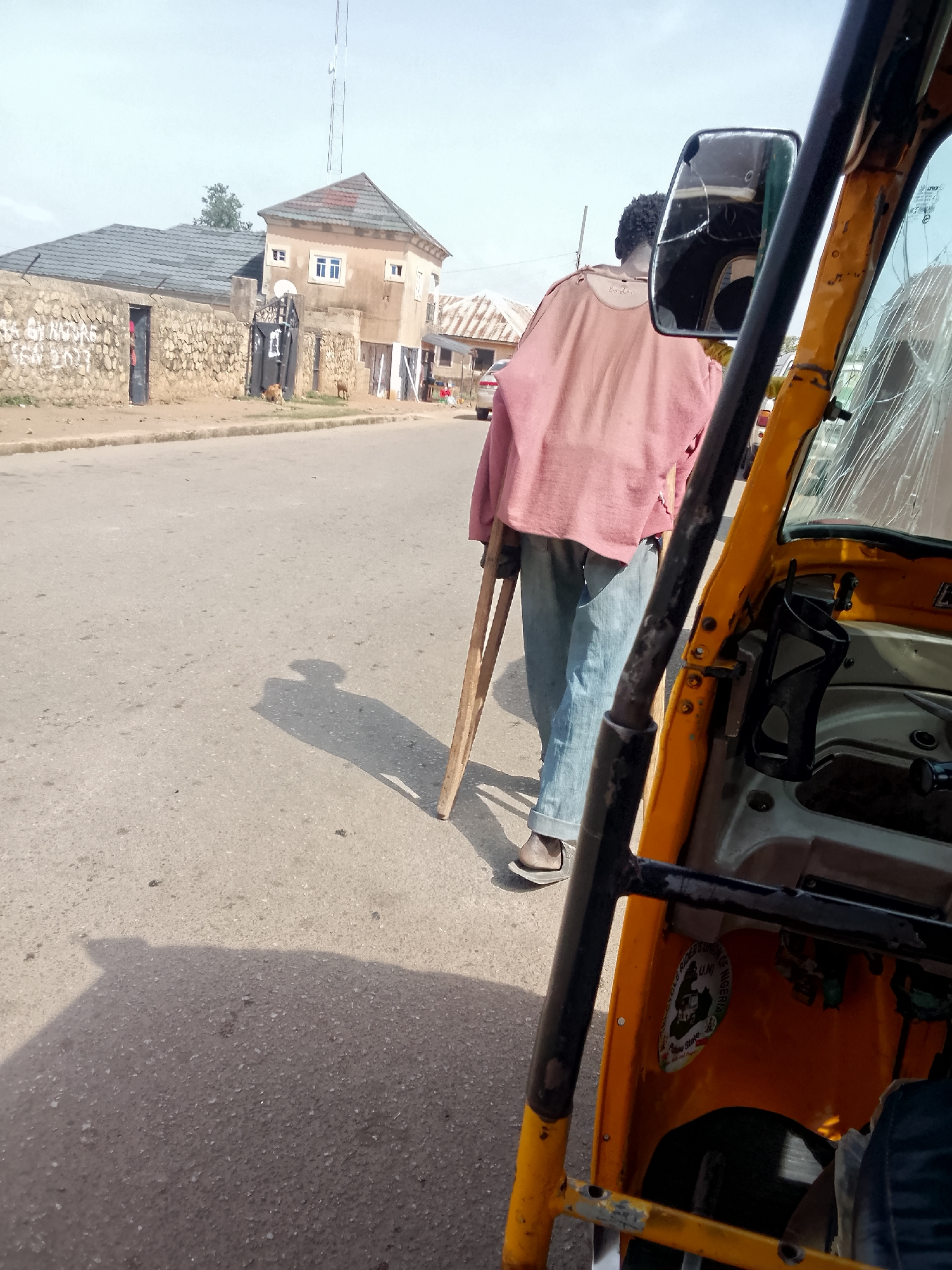 The image, taken in Bukuru, Nigeria, depicts a person with crutches walking on a paved road alongside a yellow tricycle taxi (Keke). The taxi’s windshield is cracked and a sticker is visible inside. Across the street is a stone wall in front of a two-story house with a water tower. Two dogs are at the gate. The sky is bright.