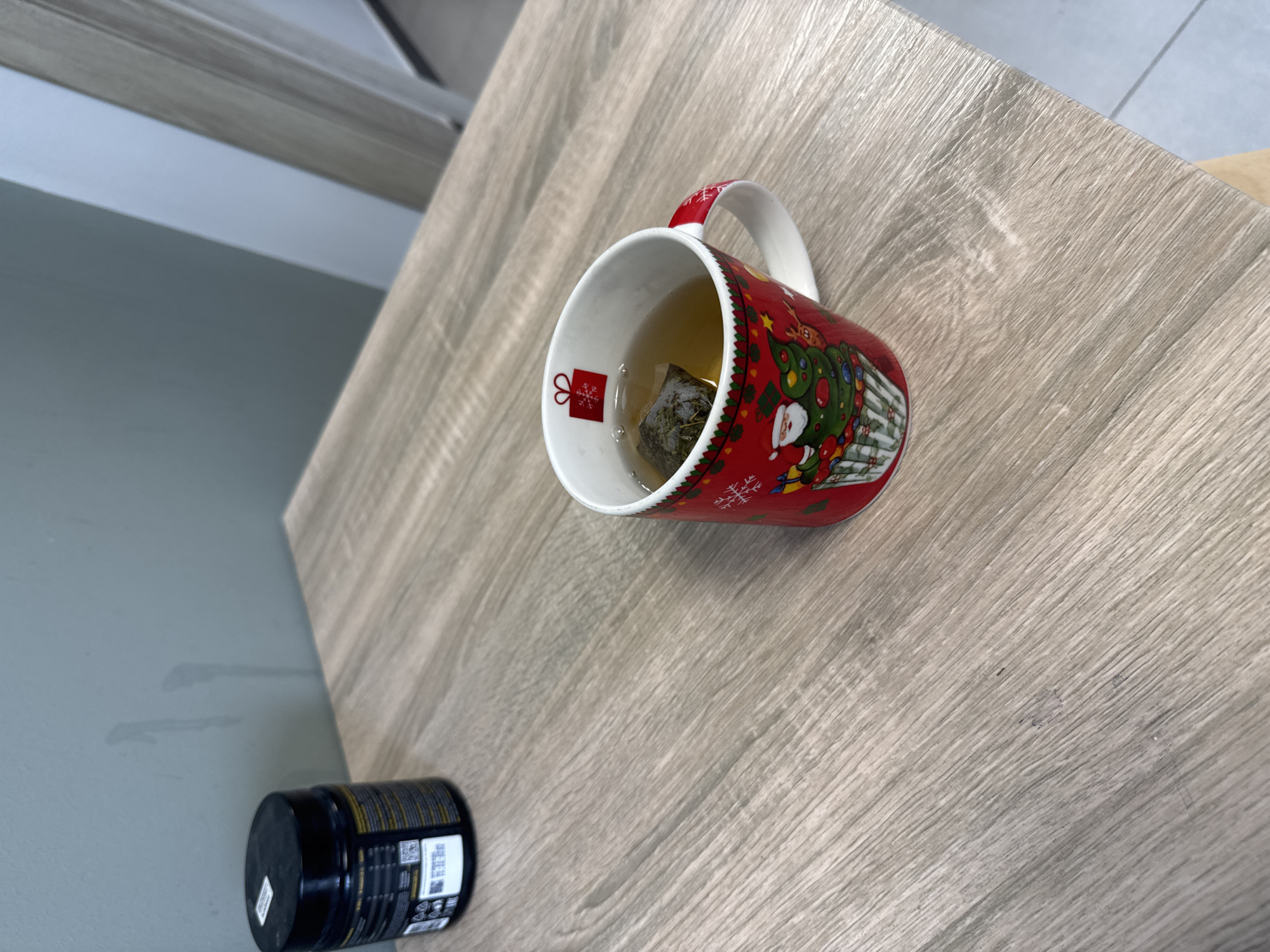 A red and white Christmas-themed mug filled with tea and a teabag sits on a light wood-grain patterned table. A black cylindrical container is situated near the bottom left corner of the image. The location is possibly a room in the city of Kazan, Russia.