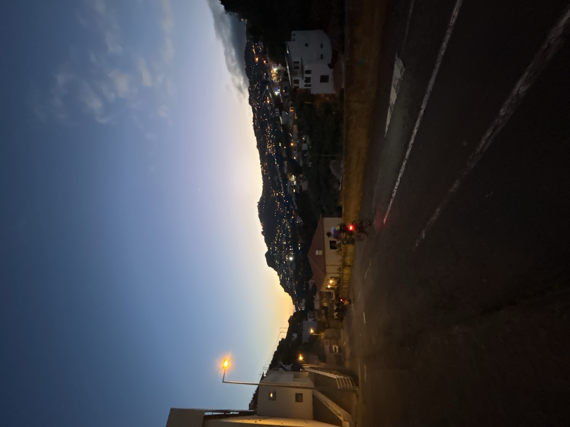 The image captures an urban scene in Câmara de Lobos, Portugal, during twilight. A hillside is dotted with residential buildings, illuminated by streetlights and house windows. The sky transitions from deep blue to orange. A two-lane road runs along the side of the image, showing the edge of a roadway with visible line markings. Two motorcyclists ride down the right side of the road. In the lower left of the frame, there is the top of a building.
