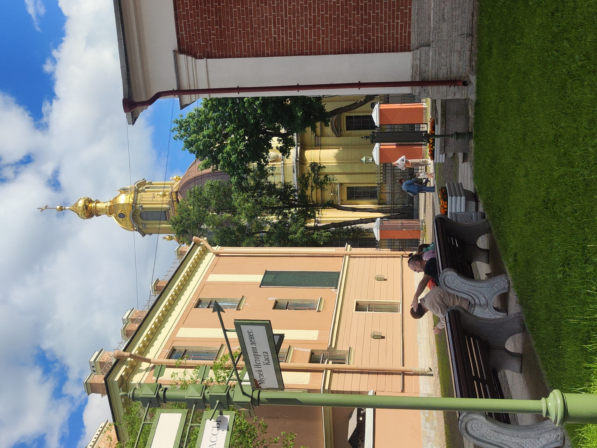 In Saint Petersburg, Russia, on a sunny day, a man and a woman sit on a park bench, while another person stands and gestures. Behind them, a signpost indicates "Museum of Money History. Cash Desk" with an arrow pointing to the right. The background features a yellow building with a golden dome and cross, typical of Russian Orthodox churches, and a red brick building. Lush green grass and trees surround the area, with a bright blue sky dotted with white clouds above.