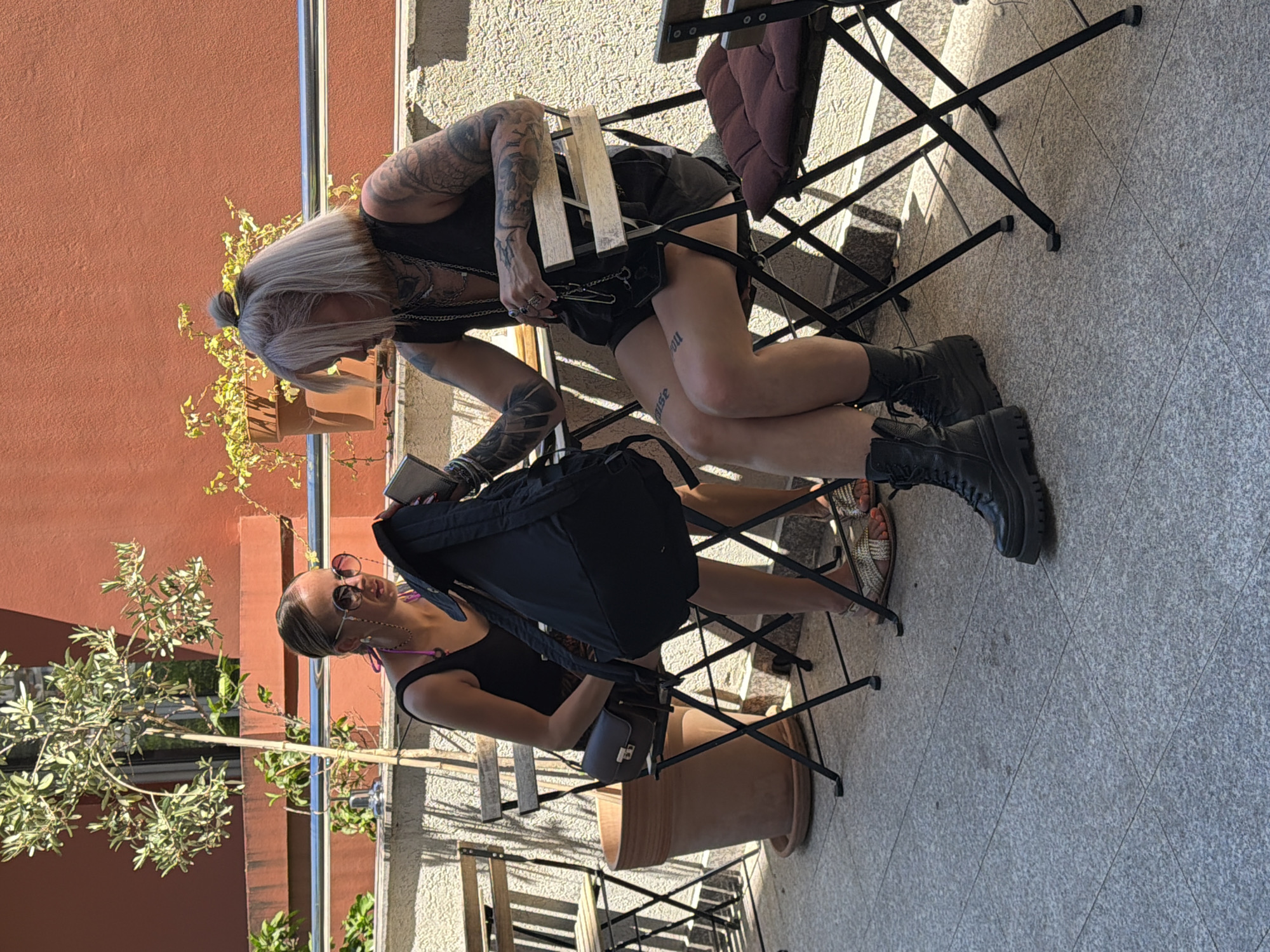 Two women are seated at separate black metal chairs on a tiled patio area. The woman on the left, with blonde hair pulled into a bun and wearing sunglasses, is dressed in a black tank top and a patterned skirt. She has a small, dark brown shoulder bag on her lap. The woman on the right has blonde hair with dark roots, tied in a ponytail, and is wearing a black tank top and black shorts. She has extensive tattoos on her arms and legs. Both women are looking down at items they are holding. The woman on the right is holding a black backpack and a small dark object. The environment includes a large terracotta pot with a small tree, another potted plant in the background, and a red wall behind the plants. The chairs are foldable, and one has a dark red cushion. The ground is paved with light grey, speckled tiles. This scene appears to be an outdoor seating area, possibly a cafe or restaurant, in Budva, Montenegro.