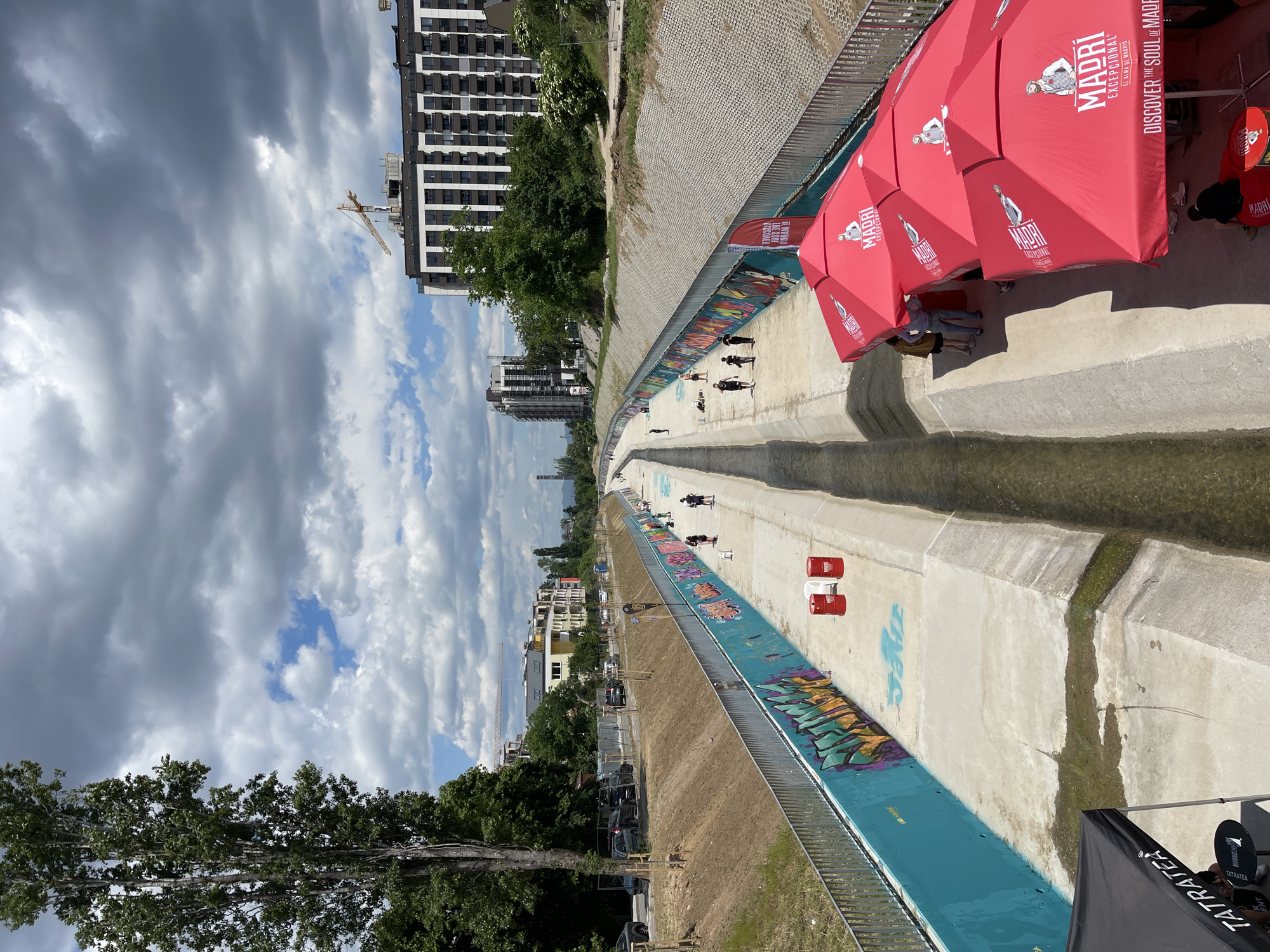 The image shows a view of a concrete canal in Sofia, Bulgaria, with red umbrellas positioned along its edge. The canal's walls are decorated with graffiti art, and people are visible walking and relaxing within the canal. Buildings of varying heights line the horizon, and a construction crane is visible among them. The sky is filled with a mix of dark storm clouds and patches of blue sky. The canal is partially filled with water. The umbrellas are branded with "MADRI Excepcional" and related advertising text.