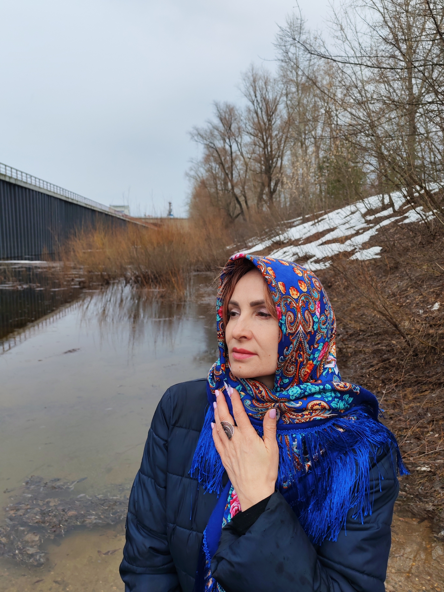 A woman stands near a body of water in Chaikovsky, Russia. She wears a blue patterned headscarf with fringe and a dark quilted jacket. Her hand, adorned with a ring, is raised to her neck. The water reflects the overcast sky and nearby bridge. Bare trees and melting snow line the shore.