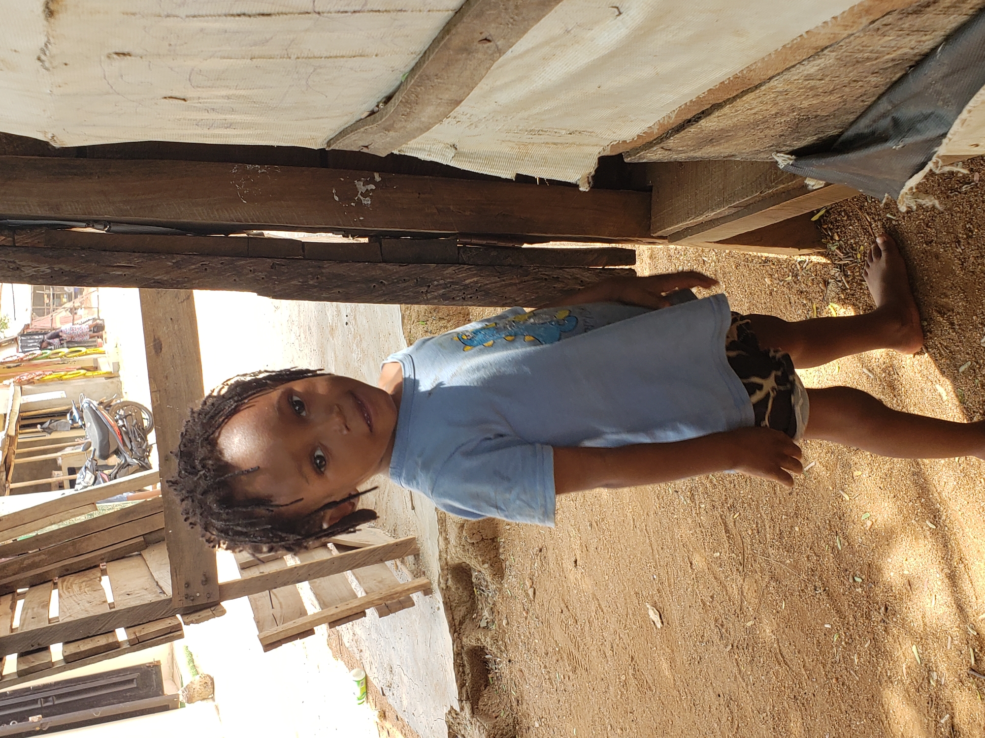 The image, taken in Adankolo, Nigeria, depicts a young child standing outdoors under bright daylight. The child, with dark skin and dreadlocked hair, is barefoot and wears a light blue t-shirt featuring a cartoon graphic and dark patterned shorts. He gazes directly at the camera with a calm, slight smile.
The setting is characterized by dry, sandy ground with scattered pebbles and plant debris. Behind the child, rustic structures are visible, constructed from wooden planks and possibly repurposed white fabric or tarpaulin used as a covering or wall. To the left, further wooden pallets or crates and part of a motorcycle or bicycle wheel can be seen, along with a white plastic bottle on the ground.
The strong illumination and clear shadows indicate a sunny day, likely midday or early afternoon. The environment suggests an informal or basic dwelling area. There is no visible, decipherable text in the image.