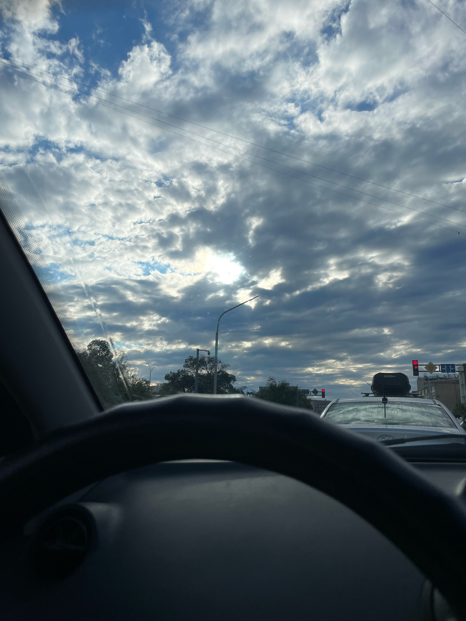 The image, taken from inside a vehicle, shows a view through the windshield of a cloudy sky over a city street. The steering wheel of the car is visible in the foreground. In the distance, there are traffic lights, street lamps, trees, and buildings. A white car with a black roof box is in front of the vehicle from which the photo is taken. The sky is dominated by dramatic clouds, with patches of blue sky visible and sunlight breaking through in some areas. The scene appears to be an urban environment in Aktau, Kazakhstan, possibly during daytime.