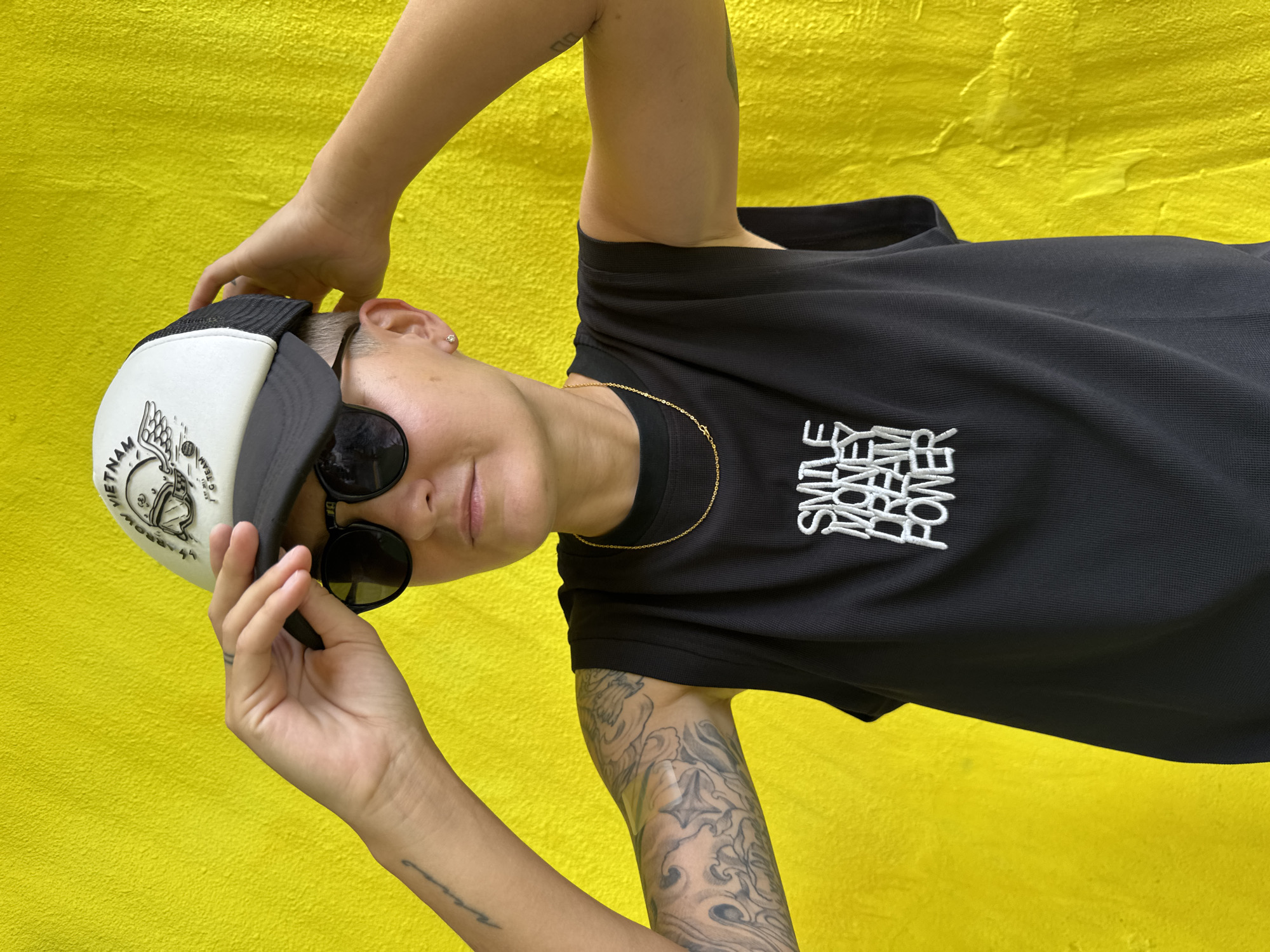 A person with short hair, wearing a black sleeveless shirt with the words "SMILE MONEY DREAM POWER" in white lettering, black sunglasses, and a black and white trucker hat with a graphic on the front, stands against a bright yellow textured wall. The person is holding the brim of the hat with their right hand and their left arm is bent with their hand resting on the back of their head. Visible tattoos adorn the person's left arm. The scene appears to be an outdoor setting, possibly in the city of Nha Trang, Vietnam, given the presence of a hat referencing Vietnam.