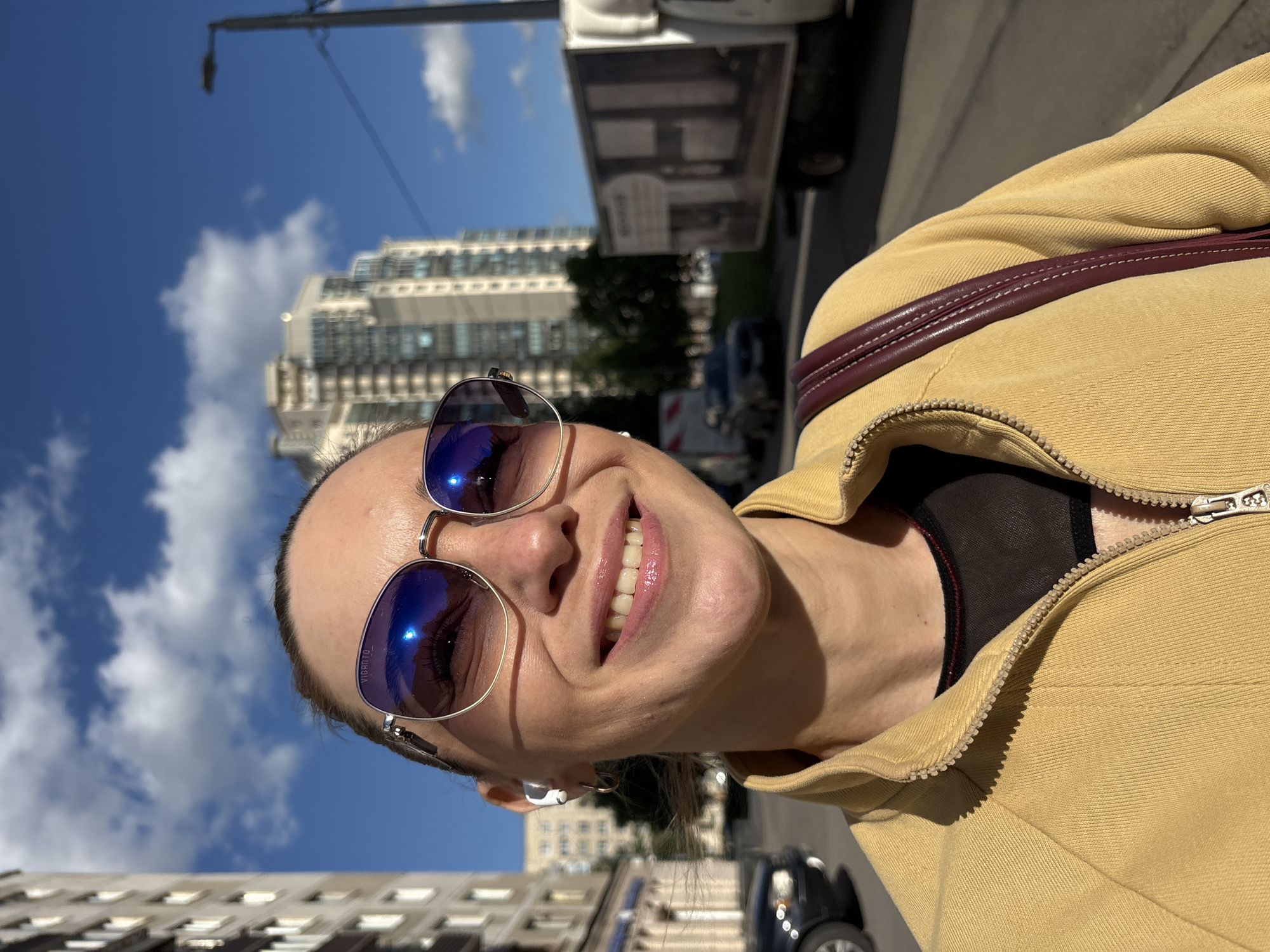 A close-up, eye-level, outdoor, selfie captures a smiling woman with blonde hair pulled back and sunglasses on, wearing a yellow zip-up jacket over a black mesh top, and a burgundy purse strap visible. She is in a city street with buildings, a truck, and cars in the background under a blue sky with clouds. The woman's left ear has a white earbud. The sunglasses have a metallic frame and the lenses reflect the blue sky and clouds. The woman's teeth are visible as she smiles. The image is taken during the day with bright sunlight.