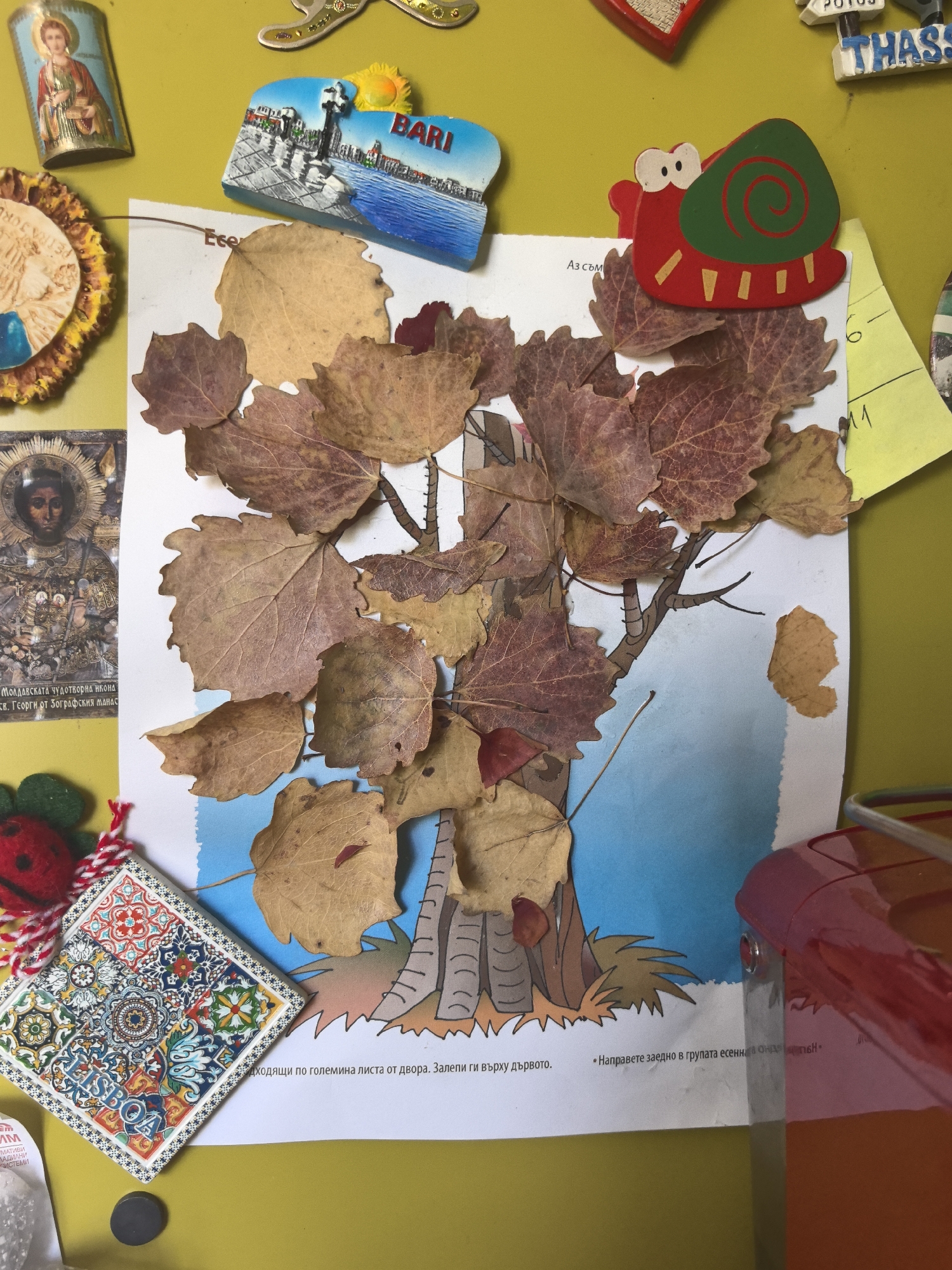 On a yellow background, likely a refrigerator door, an arrangement of objects includes religious icons, souvenirs, and a child's craft project.
The craft project is a white piece of paper with a printed illustration of a tree. Real dried leaves are glued onto the branches of the tree. The paper has text written in Bulgarian.
Around the paper are several refrigerator magnets. These include:
- Two religious icons, depicting holy figures.
- A tourist souvenir magnet from Bari, Italy.
- A tourist souvenir magnet with "THAS" written on it.
- A wooden magnet in the shape of a snail.
- A magnet made of a colorful tile with "LISBOA" (Lisbon) written on it.
- A ladybug-shaped magnet.
- A yellow sticky note.
A red plastic object is in the lower right corner, partially obscuring the scene. The location is likely Hrabrino, Bulgaria, due to the Bulgarian text on the paper.