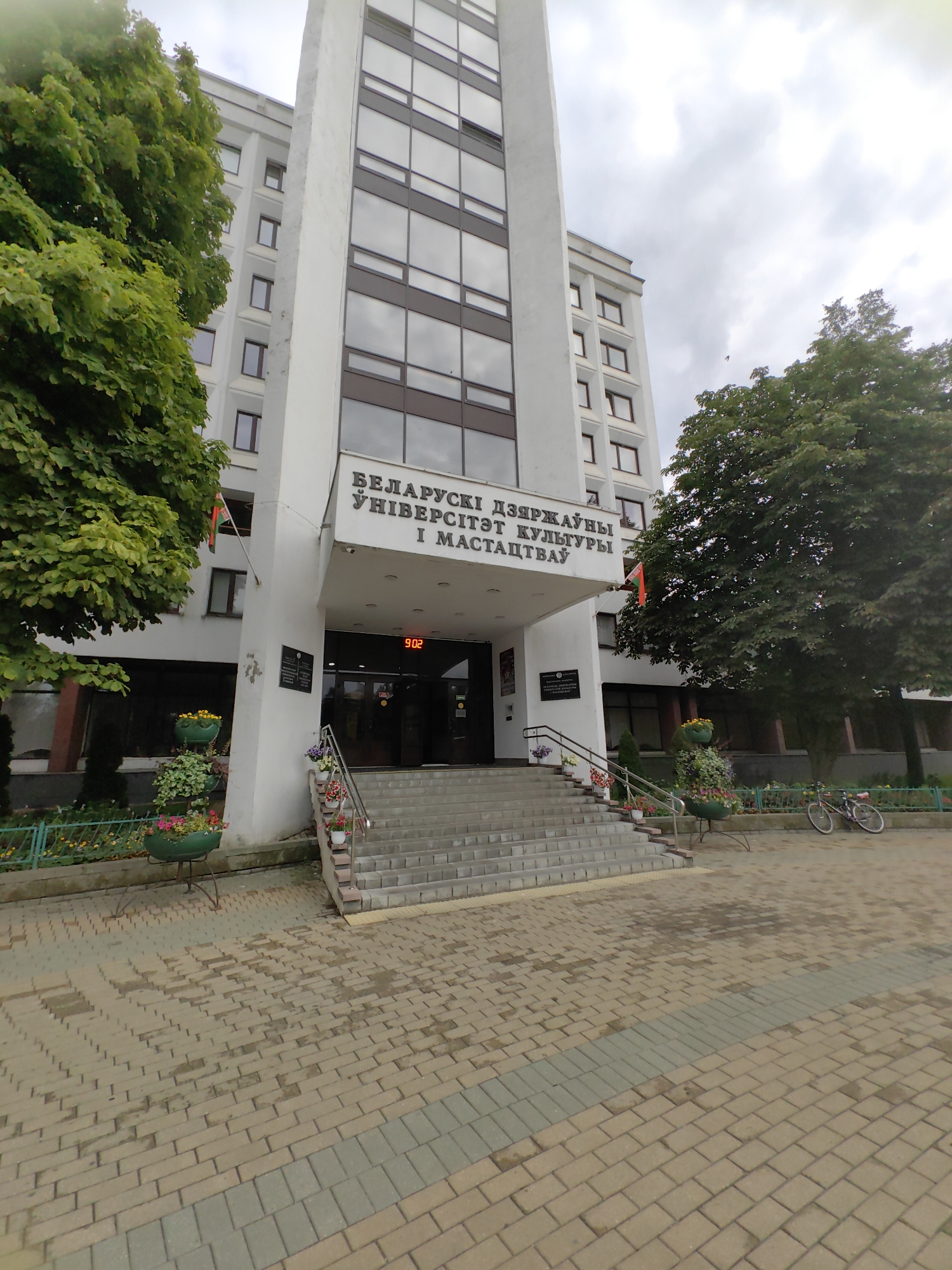 This is a photograph of the Belarusian State University of Culture and Arts in Minsk, Belarus. The building is a tall, modern structure with a white facade and numerous windows. In front of the building, there are wide steps leading up to the entrance. Flower pots with colorful flowers are placed along the steps and on stands to the left of the entrance, adding a touch of greenery and color to the scene. The entrance itself is recessed, and above it, a sign with text in Belarusian is visible. A digital clock displaying "902" is mounted above the entrance.
To the left of the building, a large green tree partially obscures the view of the facade, its leaves lush and full. A Belarusian flag is also seen hanging from a pole on the building's exterior. To the right of the entrance area, another large green tree stands. On the paved walkway in front of the building, two bicycles are parked. The ground is paved with interlocking bricks in various shades of brown and grey, and a section of the pavement has a wet appearance, suggesting it might have recently rained or been cleaned. The sky is overcast with clouds, indicating a cloudy day. The overall impression is a well-maintained public building in a city setting. The text visible on the sign above the entrance reads "БЕЛАРУСКI ДЗЯРЖАЎНЫ ЎНІВЕРСІТЭТ КУЛЬТУРЫ І МАСТАЦТВАЎ".
