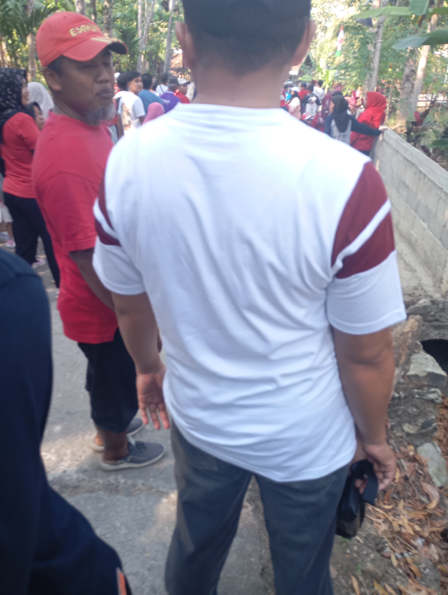 In Ketepung, Indonesia, a group of individuals are gathered outdoors. In the foreground, a person wearing a white t-shirt with maroon stripes is seen from behind, holding a black bag in their right hand. To their left, another person is partially visible, wearing a dark blue garment. Further to the left and slightly behind, a man in a red t-shirt and a red cap with "ESPANYOL" written on it is looking towards the left, with a grey beard. He is standing next to a woman wearing a white headscarf and a red shirt. In the background, other people are visible, some wearing red and white clothing, suggesting a communal gathering or event. The environment appears to be a path or walkway bordered by trees and vegetation, with a concrete structure on the right side of the image.
