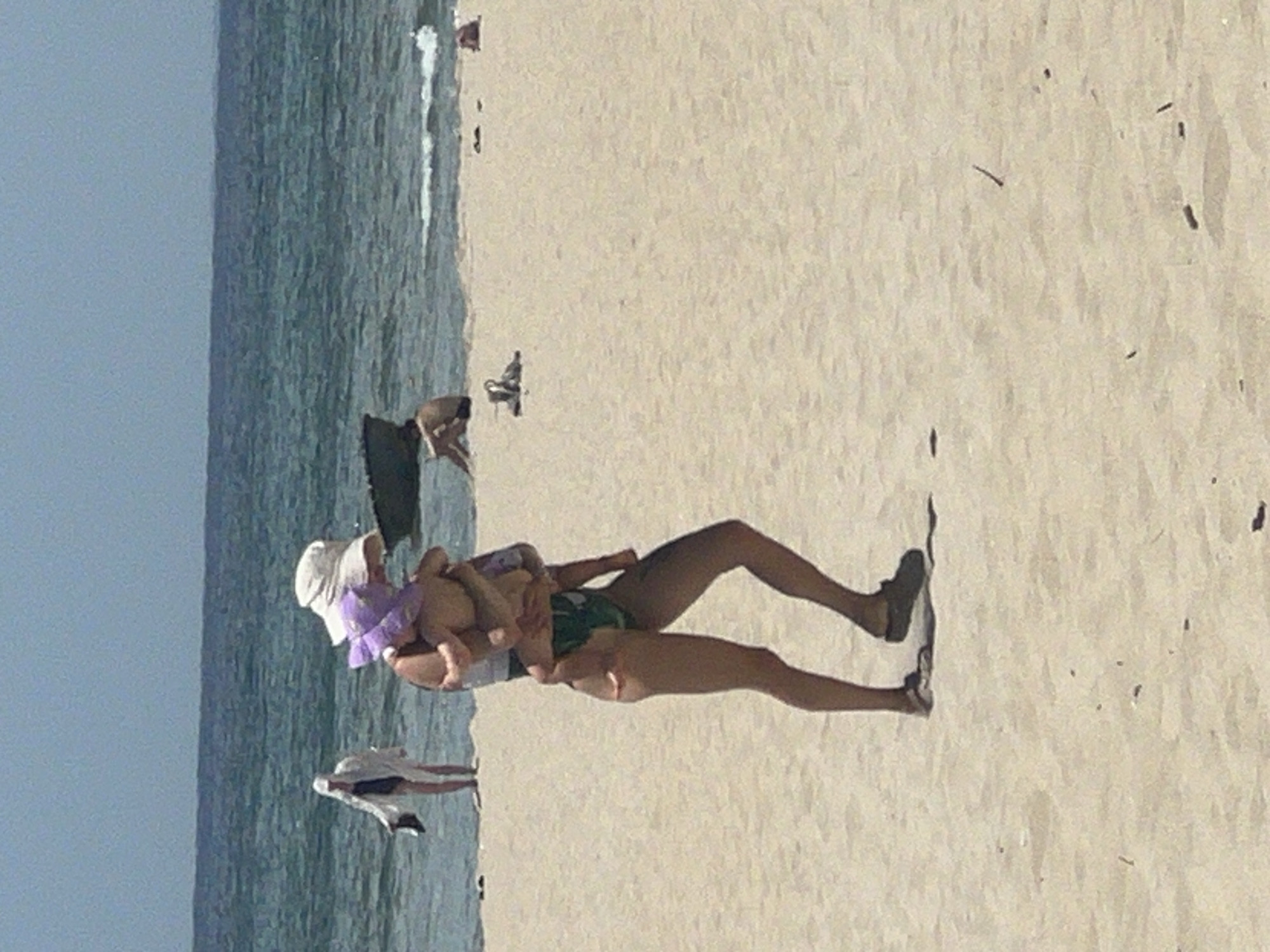 A woman is walking on a sandy beach, carrying a baby on her shoulders. The woman is wearing a white sun hat, a white top, and green and black patterned bikini bottoms. The baby is wearing a purple hat. In the background, the ocean is visible, with waves and a clear sky. Another person, possibly a woman wearing a white robe, is standing near the water's edge. There are other people in the distance, some sitting on the sand. The beach appears to be in Đà Nẵng, Vietnam.