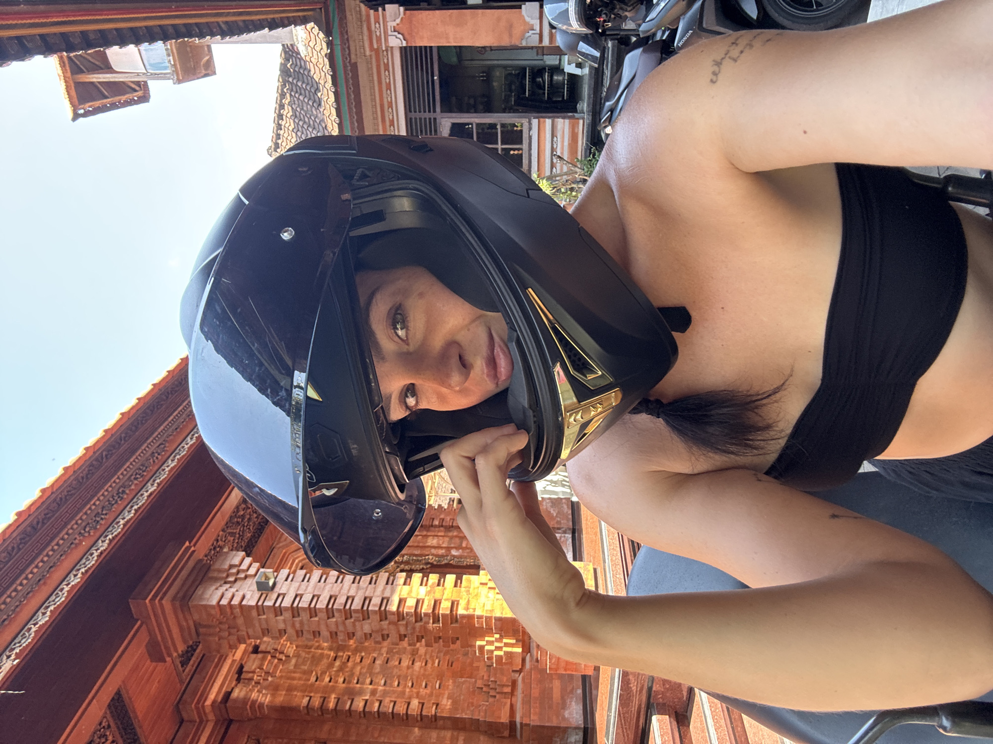 This is a self-portrait of a young woman wearing a black full-face motorcycle helmet. She is holding the helmet with her right hand, with her face visible through the visor. Her left arm is bent, and her shoulder and upper chest are exposed, wearing a black bandeau top. A dark braid of hair hangs down her back.
The background shows traditional Balinese architecture with intricately carved reddish-brown wood and tiled roofs. Sunlight is visible, suggesting it is daytime. A motorcycle is partially visible to the right of the woman. The overall impression is a casual, perhaps adventurous moment. There is text on the woman's arm, which appears to be a tattoo, but it is not clearly legible. There is also text on the motorcycle which reads "HONDA". The scene suggests a location in North Kuta, Indonesia.