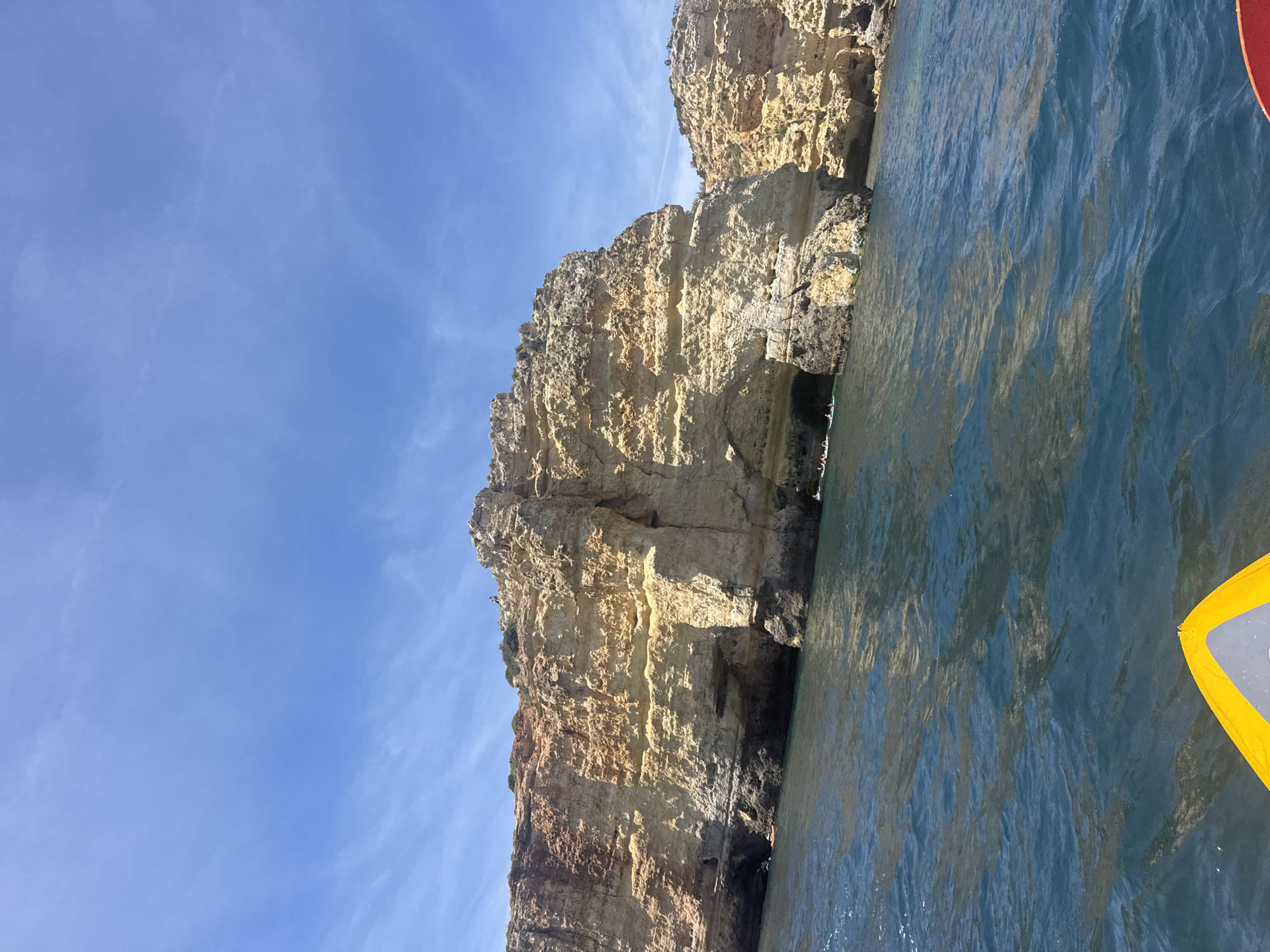 Here's a detailed description of the image:
**Setting:** The image depicts a coastal scene with a large, imposing cliff face meeting the water. The location is unknown, as the city is listed as "Unknown."
**Cliff:** The cliff is composed of layered rock formations, showing a mix of light yellow, tan, and darker brown hues. The cliff's texture is rugged and uneven, with some areas appearing smoother than others. Several small cave-like openings can be seen at the cliff's base.
**Water:** The water is a deep blue color, reflecting the sky. It has a rippled surface, indicating movement and light reflecting off the waves.
**Sky:** The sky is a clear blue with scattered white wisps of clouds.
**Foreground:** In the lower right corner of the image, part of a yellow and grey object is visible, possibly part of a boat or watercraft.
**Overall Impression:** The scene is a natural landscape highlighting the dramatic interaction between the sea and the cliffs. The bright sky and the textures of the rock formations create a visually striking image.