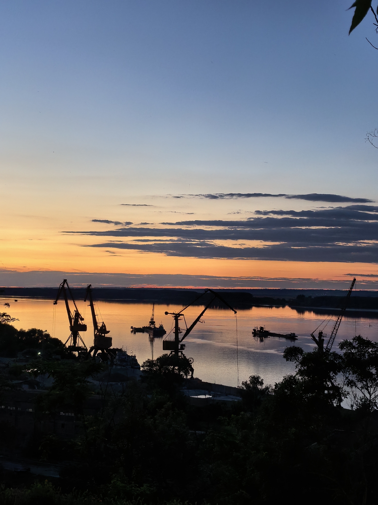 This is a photograph of a river port at dusk in Svishtov, Bulgaria. The sky is a gradient of blue at the top, transitioning to orange and yellow near the horizon, with some scattered dark clouds. The river reflects the colors of the sky and appears calm. Several large industrial cranes are silhouetted against the sky and river in the foreground and midground. There are also several barges or boats on the water. The foreground is dark and appears to be a hillside covered in trees and vegetation, obscuring much of the immediate port infrastructure on the left. The overall scene depicts a working port at the end of the day, with the natural beauty of the sunset creating a dramatic backdrop.