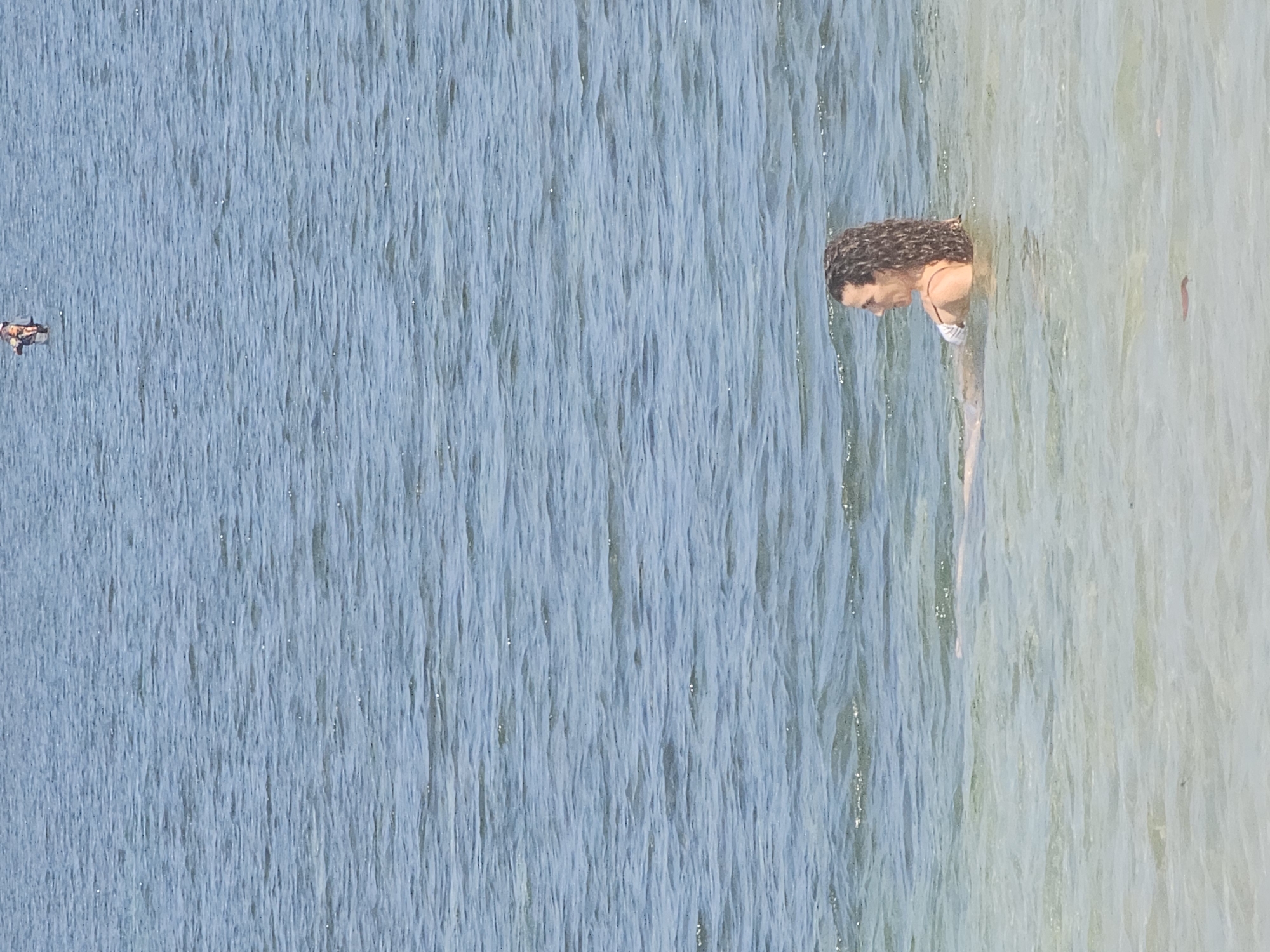 A woman with curly brown hair is swimming in a body of water, wearing a white bikini. Her body is partially submerged, and she is facing to the left. In the distance, a person is visible standing in the water. The water is blue and appears to be calm with small ripples. The scene is set outdoors.