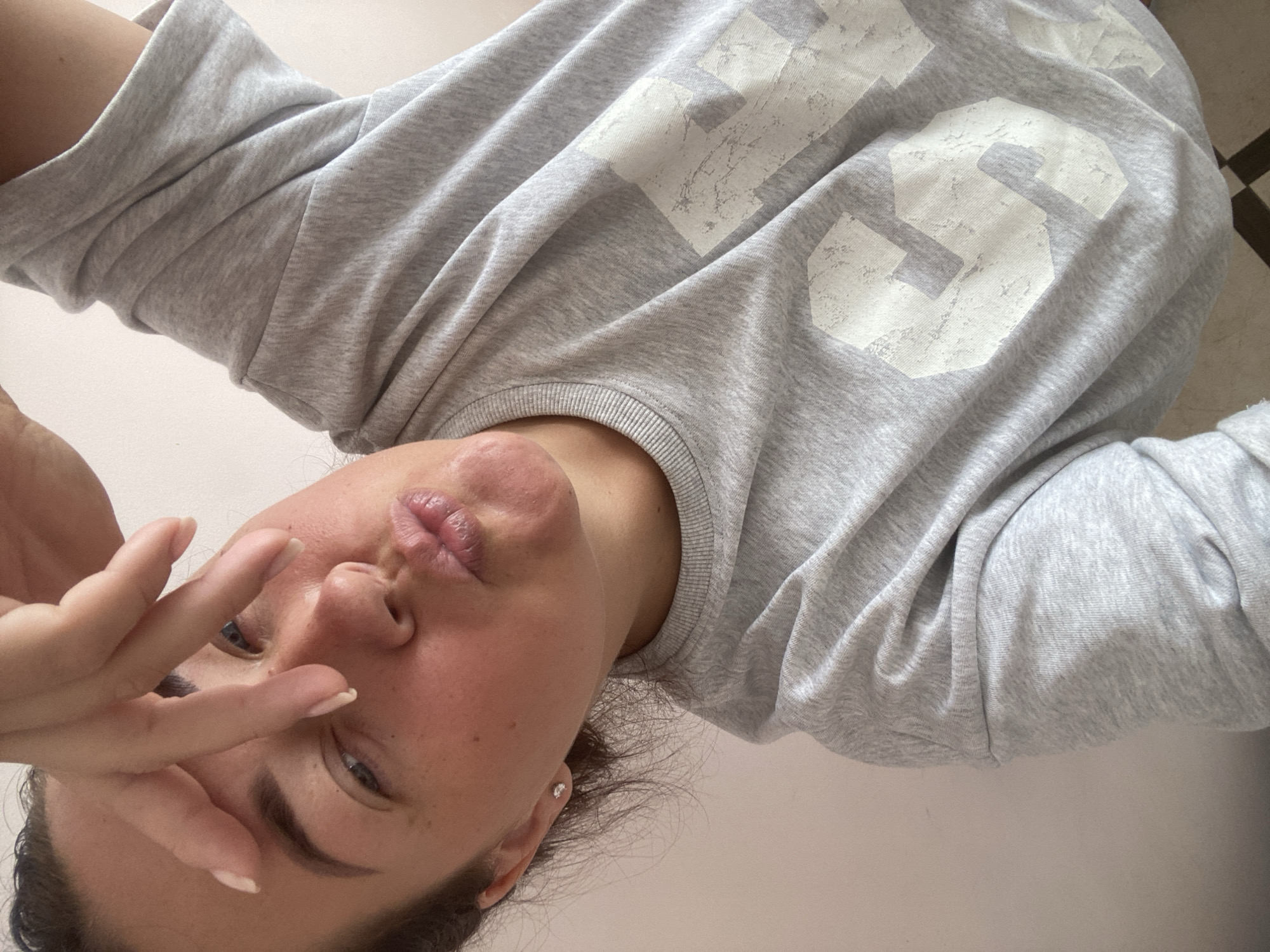 The image is a close-up, low-angle selfie of a young woman with medium-toned skin, freckles, and dark hair pulled back. She is wearing a light gray sweatshirt with a faded, large white graphic of the number "34" on it. Her right hand is raised to her forehead, obscuring part of her face. Her lips are pursed. The background is a plain, light-colored wall. The flooring visible in the bottom left corner appears to be tiled in a checkerboard pattern. The lighting suggests an indoor setting. There is no identifiable information to indicate the location is in Sochi, Russia.