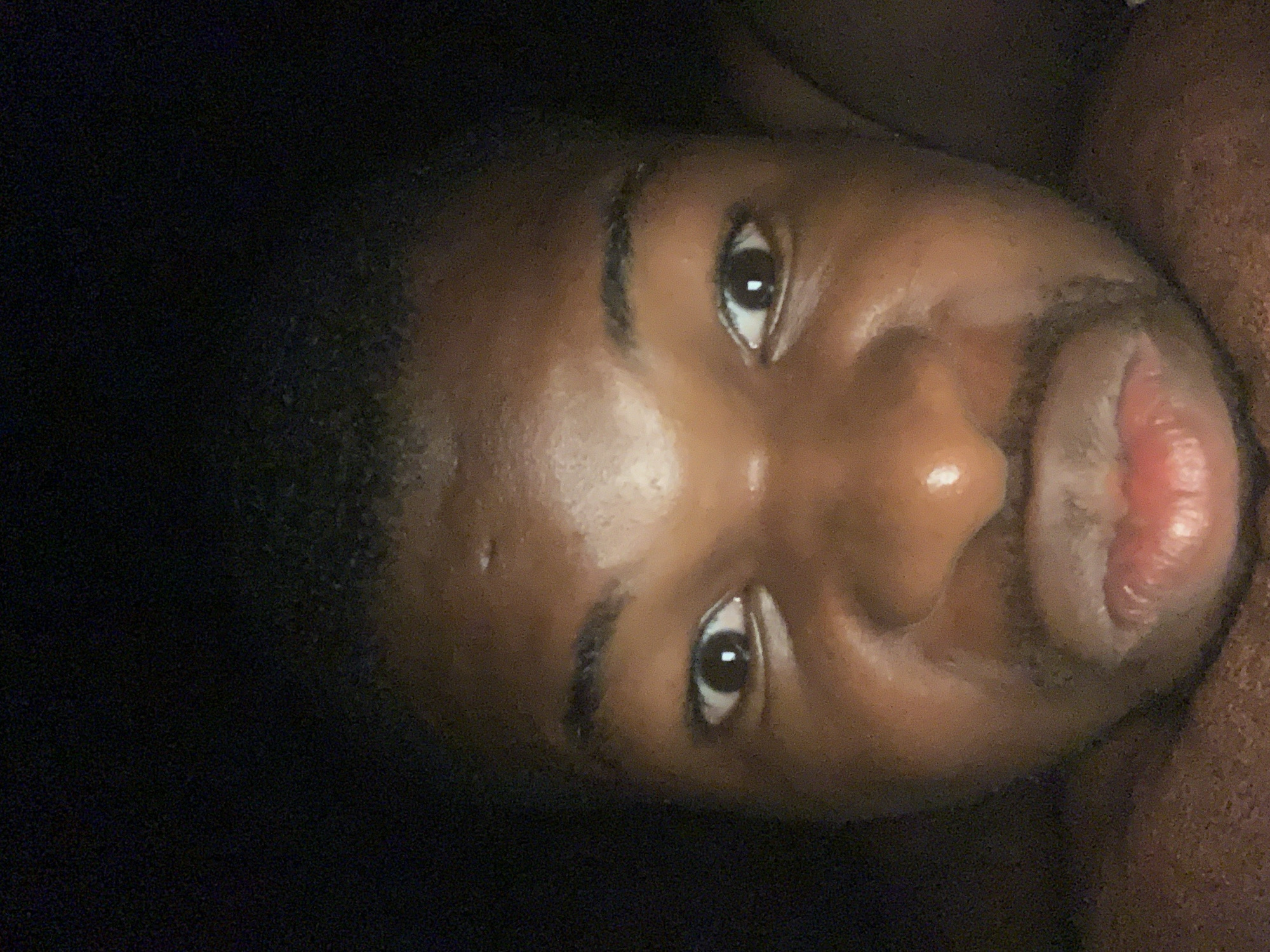 A close-up, slightly tilted portrait of a dark-skinned man with short hair, dark eyes, and dark eyebrows. His lips are slightly parted. The background is mostly dark and out of focus. There is no information in the image to suggest this was taken in Onitsha, Nigeria.