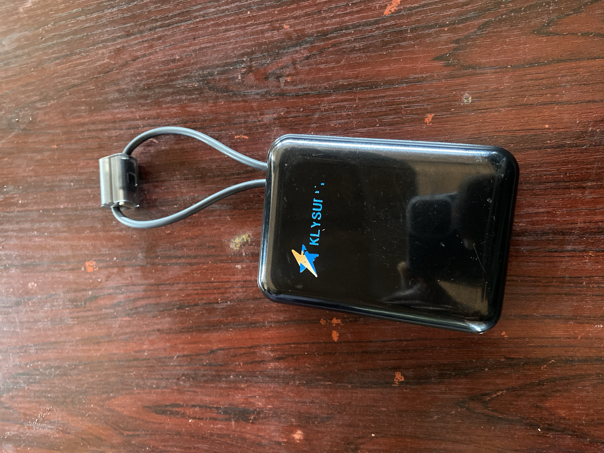 A black, rectangular electronic device with rounded corners rests on a reddish-brown wooden surface, located in the city of Howayi, Nigeria. The device features a glossy finish and displays a blue and yellow/orange stylized lightning bolt or star logo, followed by the blue text "KLYSUI". A grey, flexible strap or cable emerges from the top of the device, forming a loop secured by a silver-toned cylindrical metal clasp positioned at its apex. The wooden surface exhibits prominent vertical grain patterns and minor surface wear, including faint scratches and localized lighter discolorations. No individuals or active interactions are visible in the scene.