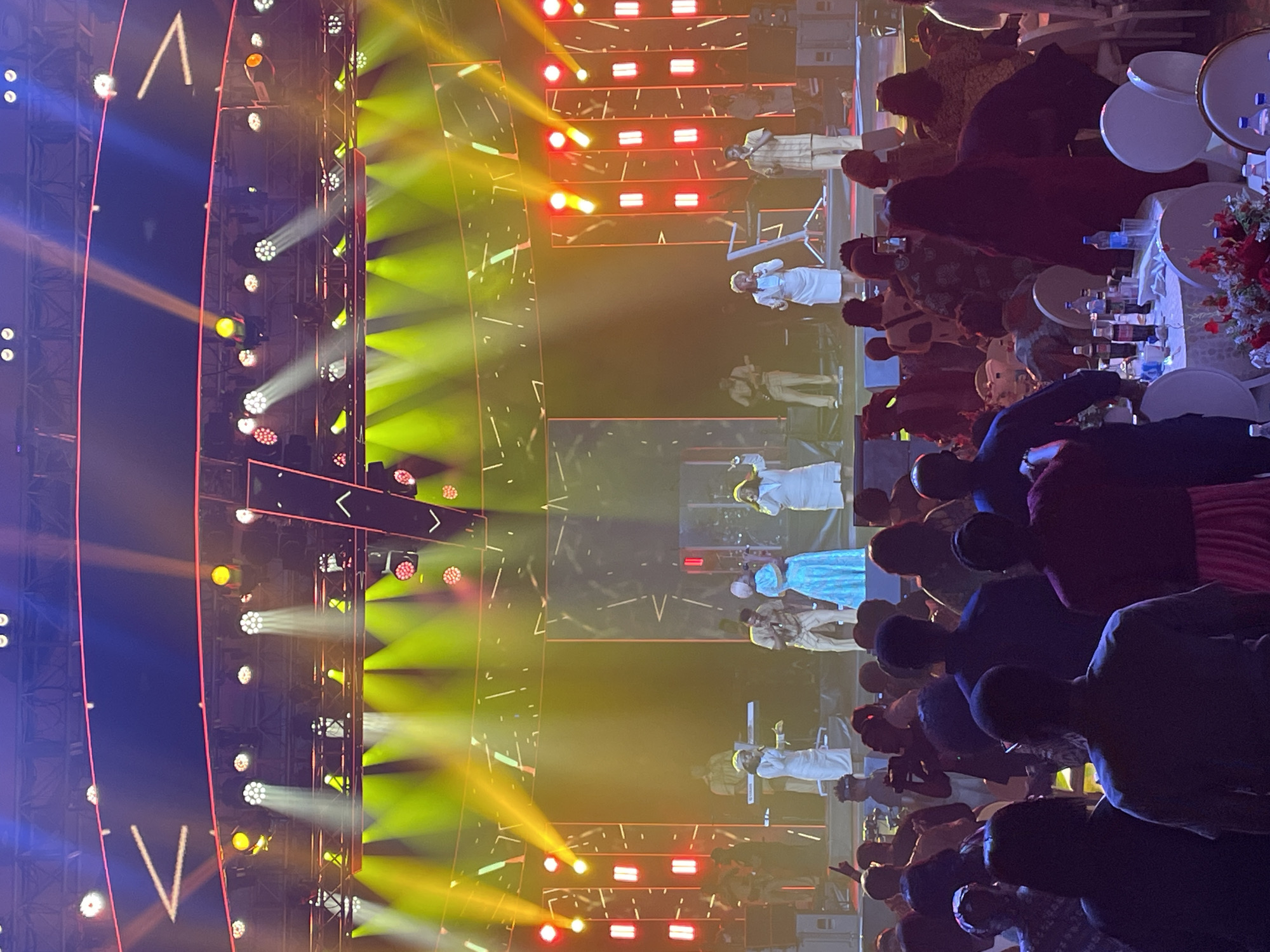 The image shows a performance on stage in Itirin, Nigeria. The stage is brightly lit with spotlights of white, yellow, and red. There are multiple performers dressed in various colors, including white and blue. The audience is visible in the foreground, seated at tables with white tablecloths, bottles, and flower arrangements. Some audience members appear to be recording the performance with their phones. The stage backdrop consists of a structure with lights.
