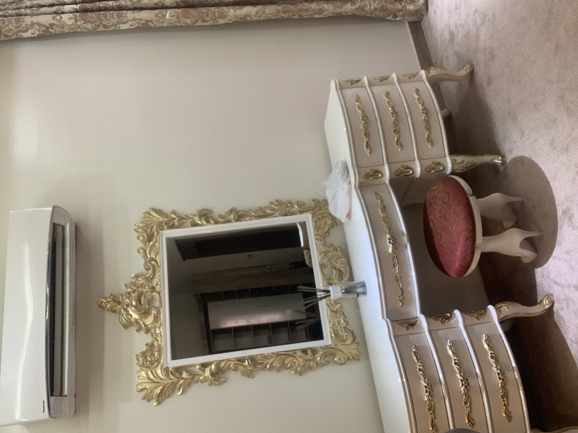 The image depicts a bedroom scene in Abuja, Nigeria. The primary subjects are a vanity table with drawers and a matching stool, a large ornate mirror with a gold frame, and an air conditioning unit mounted on the wall. The vanity and stool are a cream color with gold detailing on the drawer fronts and legs, exuding a luxurious and classic aesthetic. The stool is upholstered in a rich red fabric with a patterned texture. The mirror's frame is elaborately carved with ornate gold swirls and flourishes, reflecting the interior of the room. The air conditioning unit is a sleek white rectangle.
The scene is brightly lit, suggesting it is daytime, and there are no indications of specific weather conditions. The overall setting appears to be a well-furnished and well-maintained room, likely a master bedroom or dressing area, given the presence of the vanity and mirror. The soft carpeting suggests a comfortable indoor environment. There is no activity or event taking place in the image; it is a static shot of the room's furnishings.
Notable details include the intricate gold ornamentation on the furniture and mirror, which are characteristic of a classical or baroque style. The reflection in the mirror shows more of the room, including shelving units, suggesting ample storage. The Panasonic brand name is partially visible on the air conditioning unit. The image conveys a sense of elegance and opulence.