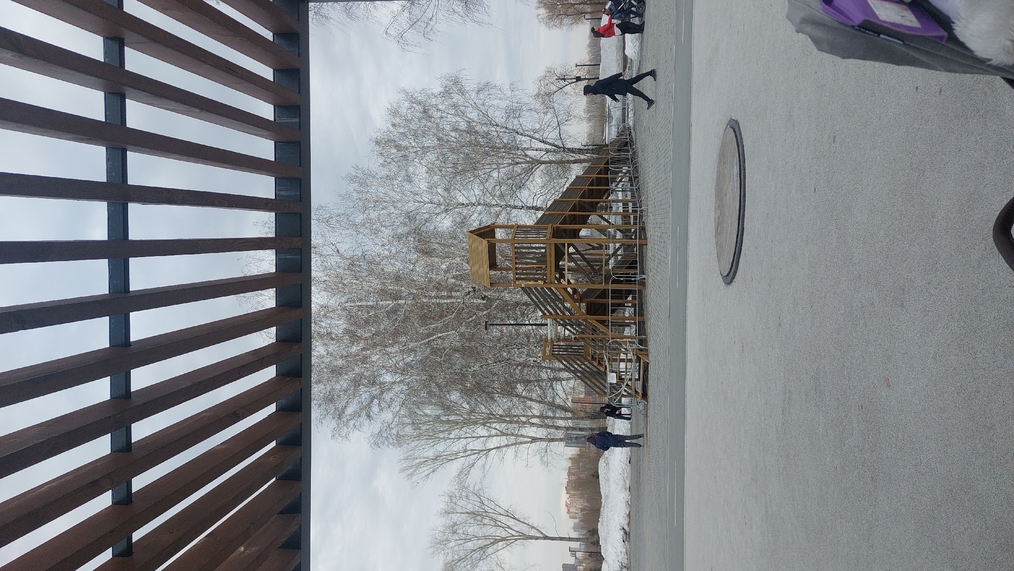 The photograph was taken in Novosibirsk, Russia, and shows an outdoor space, likely a playground or park. A tall, wooden climbing structure with multiple levels and staircases stands in the middle. Bare trees surround the structure, suggesting it is either late fall or winter. Several people are in the vicinity, including at least one person walking or running. The ground is paved with light-colored material. A partially visible wooden structure with horizontal slats is in the foreground, possibly providing shade or acting as a decorative feature.