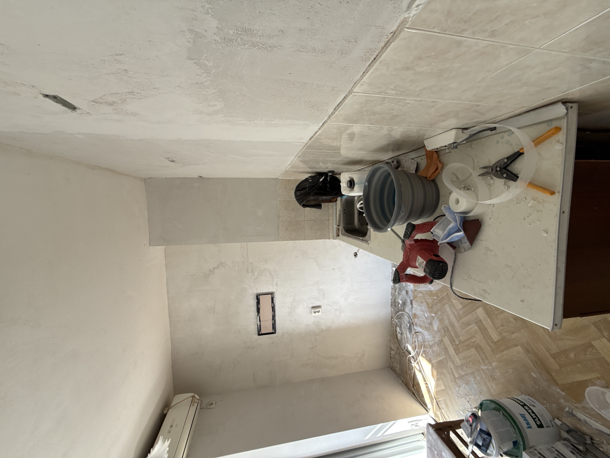 The image depicts an interior space undergoing renovation. The walls are plastered but not yet painted, and the ceiling appears to be partially finished. There's a rectangular opening in the wall, presumably for an electrical outlet or similar fixture. Beneath the window is an air conditioning unit. On the right side, there's a countertop covered with various construction-related items, including a bucket, a sander, and tools. The floor is parquet. The location is Stara Zagora, Bulgaria. No people are visible in the image.