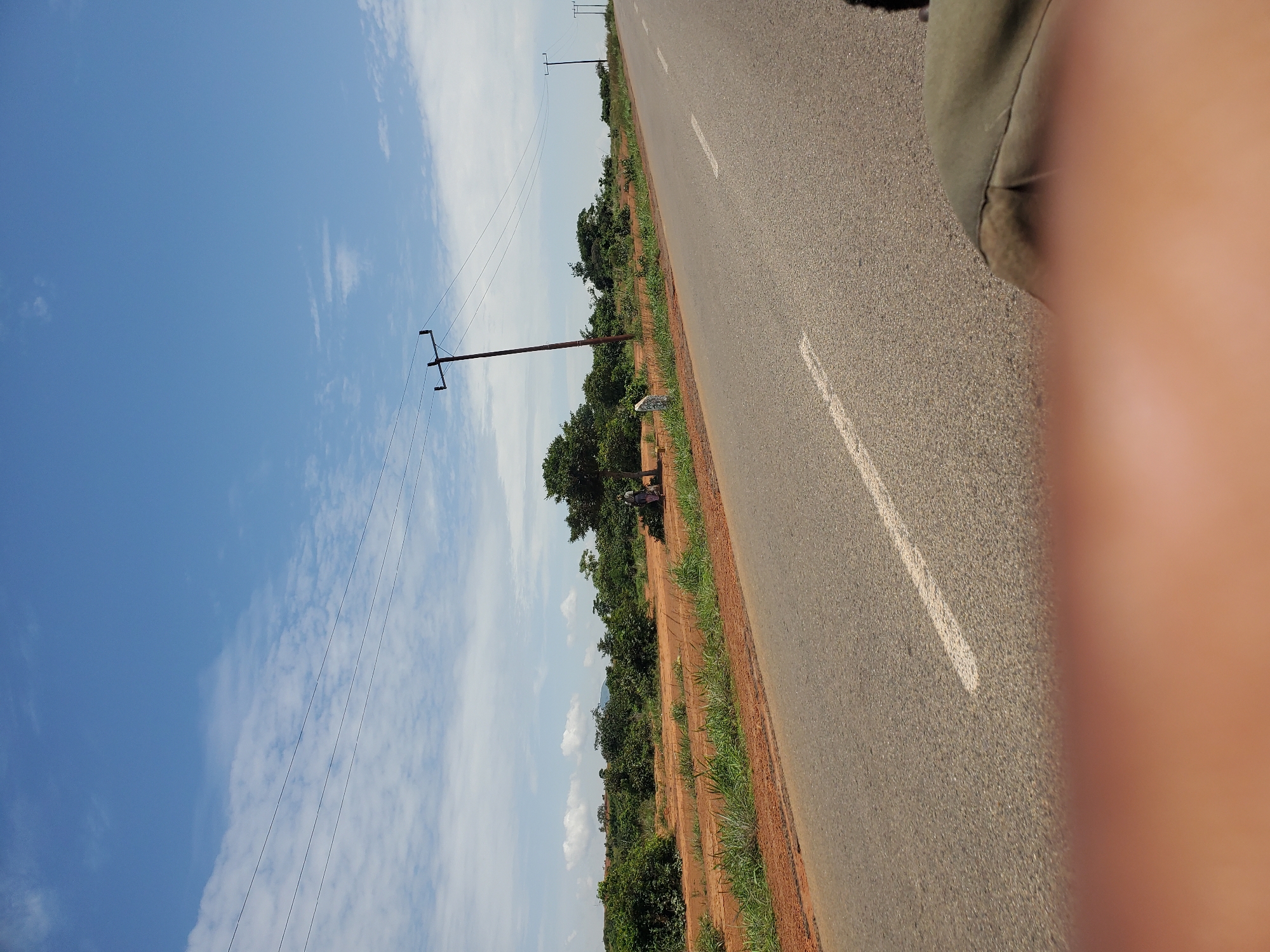 A road runs through a rural landscape in Pankshin, Nigeria. The road is paved and has a white dashed line marking the center. On the left side of the road is a dirt shoulder with sparse green grass and some shrubs. Further back are more trees and bushes. On the right side of the road, the landscape continues with similar vegetation and scattered trees. Power lines are strung across the scene, supported by wooden poles. In the distance, rolling hills can be seen under a bright blue sky with scattered white clouds. The overall scene depicts a sunny day in a rural area with evidence of electrical infrastructure.