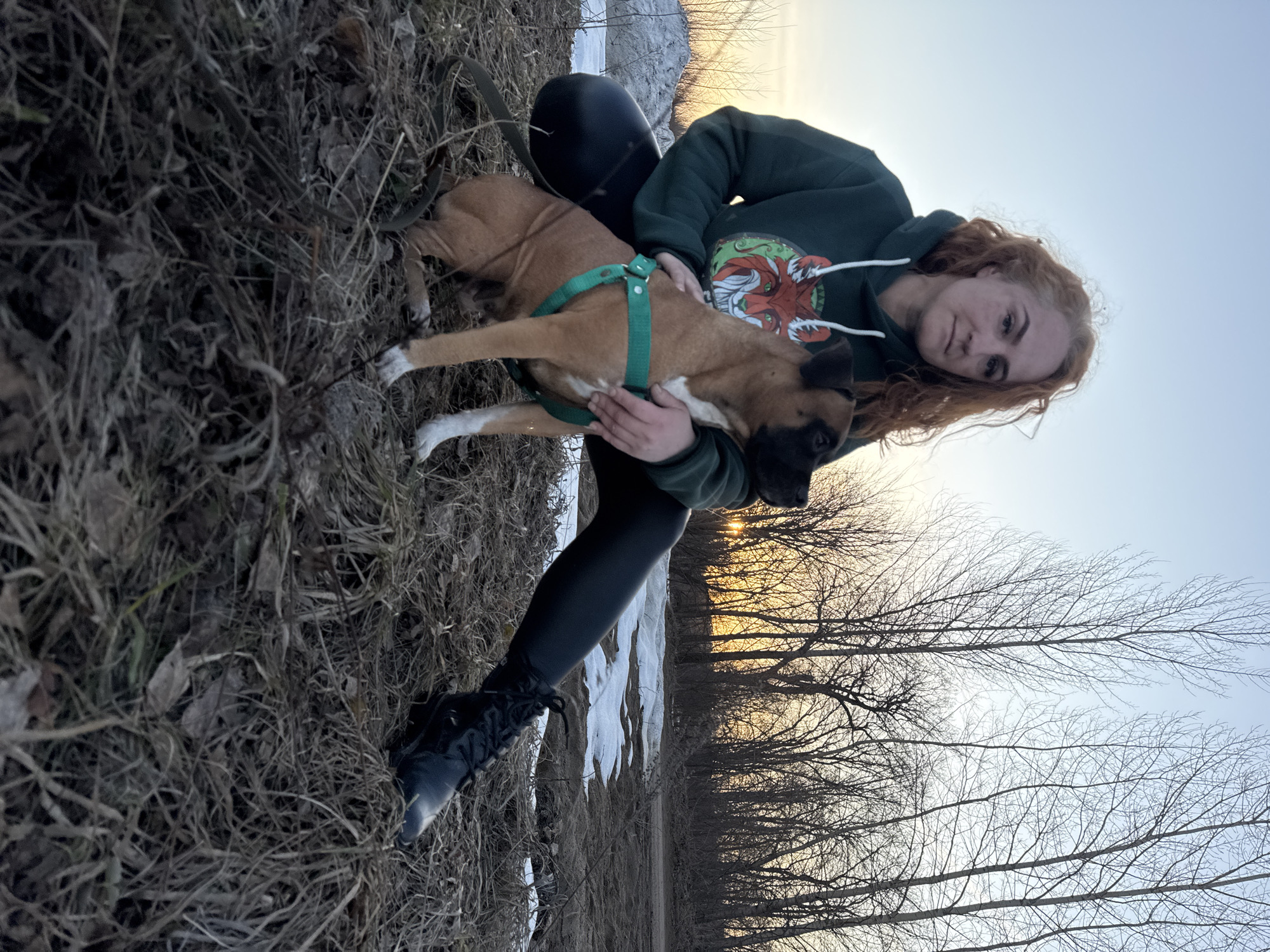 A young woman with auburn hair is hugging a tan and black dog wearing a green harness outdoors. The woman is wearing a dark green hooded sweatshirt with a graphic on the front and black leggings and boots. The dog has white markings on its legs. They are positioned near dried grass and leaves, with patches of snow visible in the background. In the distance are trees with bare branches against a light sky. There is no information in the image to identify the city as Noviy, Russia.