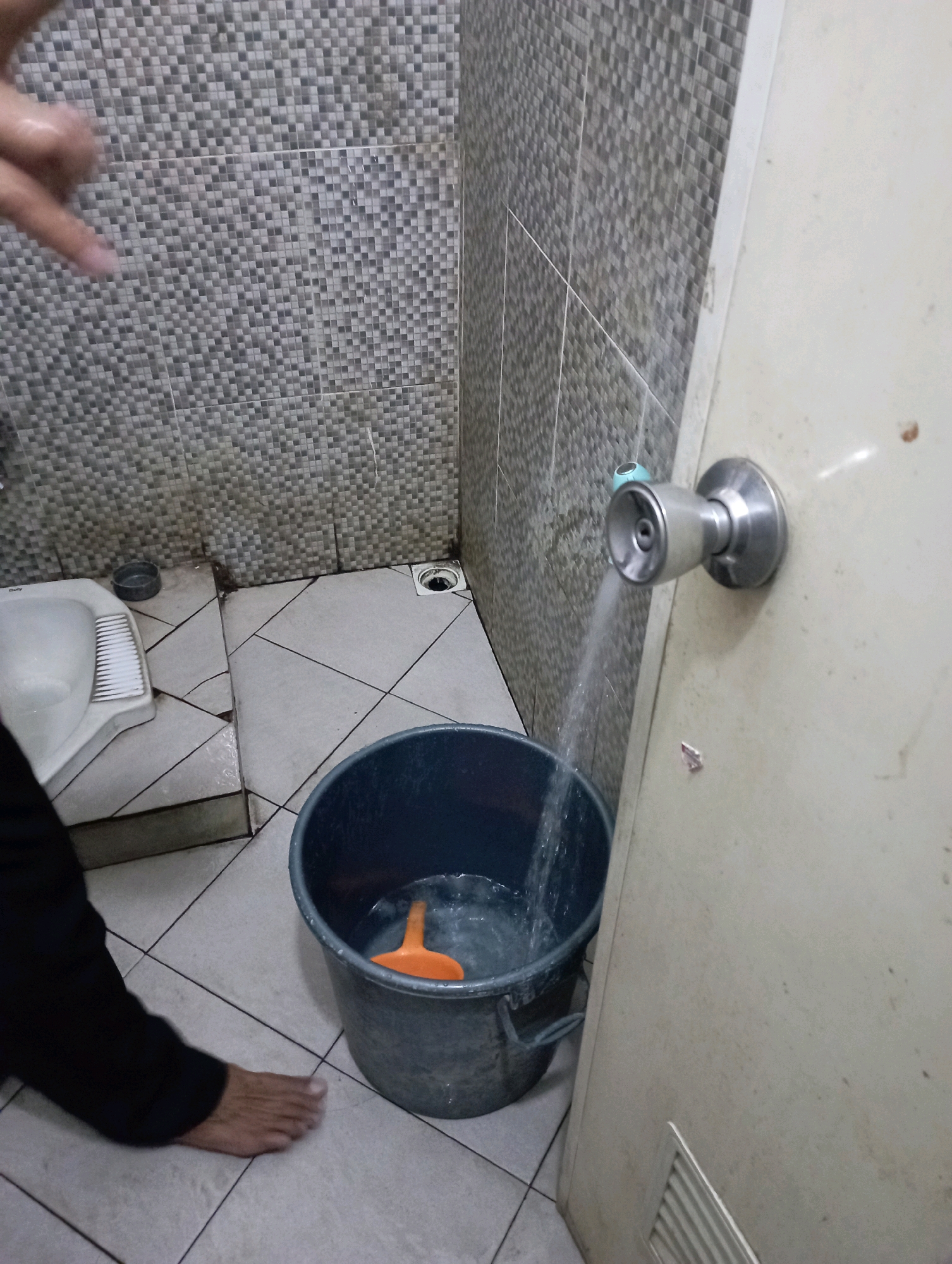 The image depicts a cramped, tiled bathroom, likely in Tasikmalaya, Indonesia, given the style of the squat toilet and the common practice of using buckets for water. Water is seen spraying from a faucet attached to a door handle, filling a large dark grey plastic bucket with an orange scoop inside. To the left of the bucket is a white squat toilet with a ridged seat. A person's bare left foot and lower leg, clad in black pants, are visible in the foreground, suggesting someone is standing near the bucket. The walls are covered in small, variegated grey and white square tiles, with a different, larger tiled pattern visible on the right wall behind the door. A drain is visible on the floor near the toilet. The overall impression is of a utilitarian and somewhat aged bathroom environment.