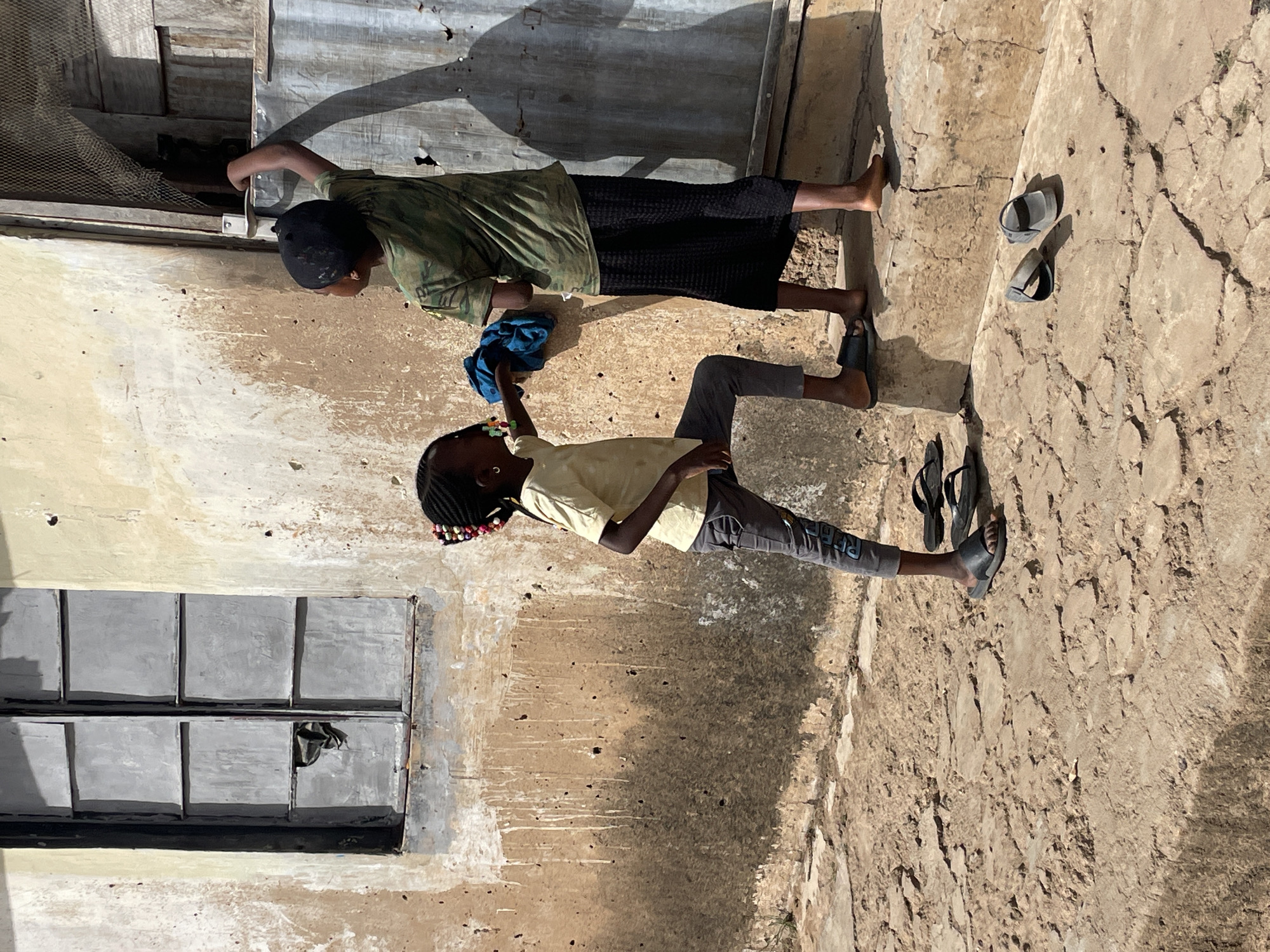 This image, captured in Jos, Nigeria, presents an outdoor scene featuring two individuals, likely children or young people, under bright daylight. The setting appears to be an urban or semi-urban area, possibly a courtyard or an alleyway between buildings.
The ground is dry, cracked, and uneven, consistent with an arid or dusty environment. On the left, a light-colored wall, possibly stucco or painted concrete, shows signs of weathering and dirt. A multi-pane window with grey, square sections is set into this wall. Towards the center and right, behind one of the subjects, is a corrugated metal structure, possibly a door or the side of a shed, casting a prominent shadow.
The taller individual, standing on the left, wears a dark cap, a green camouflage-patterned t-shirt, and a dark skirt or trousers. They are barefoot and are seen reaching towards or holding a part of the metallic structure, with a blue cloth clutched in one hand.
The shorter individual is positioned to the right, wearing a light yellow t-shirt and grey trousers with some indistinct lettering visible on the leg. Their hair is braided and decorated with colorful beads. They are wearing dark flip-flops, and additional pairs of flip-flops are scattered on the ground near their feet and further to the right. The strong shadows cast by the subjects and structures indicate a sunny day, likely during the midday hours. The overall impression is one of everyday activity in a simple, outdoor environment.
