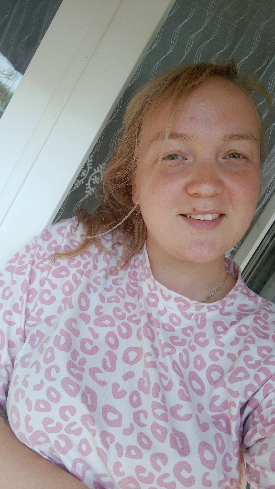 A woman with light blonde hair and fair skin is posing for a selfie. She is wearing a white shirt with pink animal print. A silver chain is visible around her neck. Behind her is a white-framed window, and a patterned grey curtain is partially visible through the window. The image may have been taken indoors. The location of Уральское, Russia cannot be determined from this image.