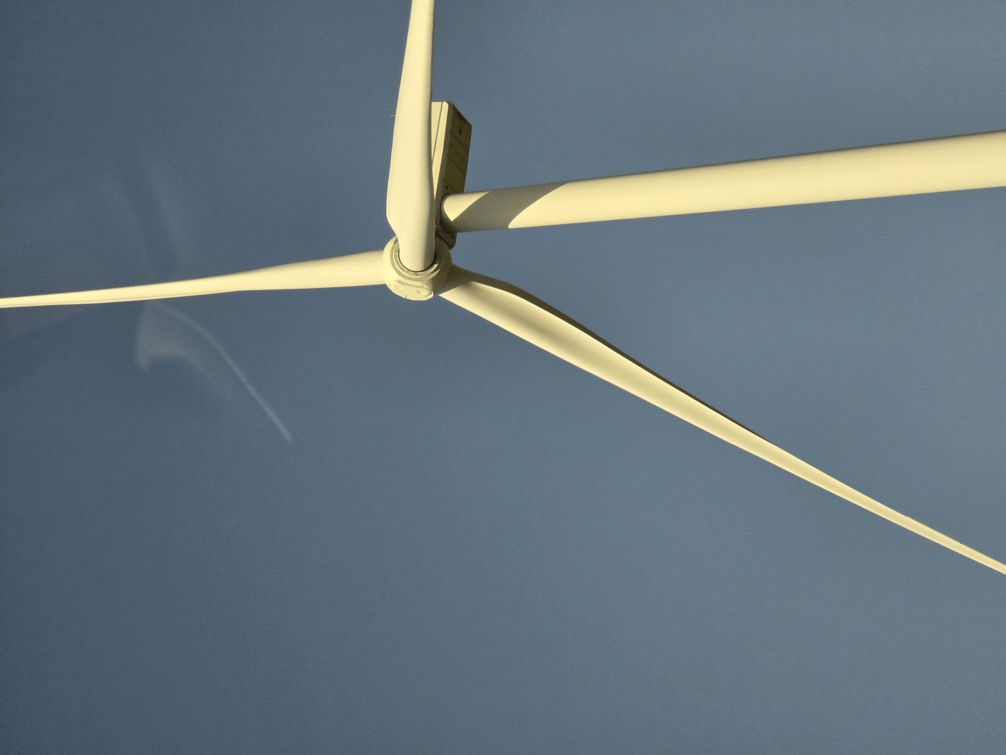 A close-up, low-angle shot focuses on a portion of a white wind turbine against a clear, blue sky. The turbine's pole and two of its blades are visible, with the blades angled upwards and to the right. A faint contrail or cloud streak is visible in the upper portion of the sky. The scene is devoid of people. This image was taken in Langeweg, Netherlands.