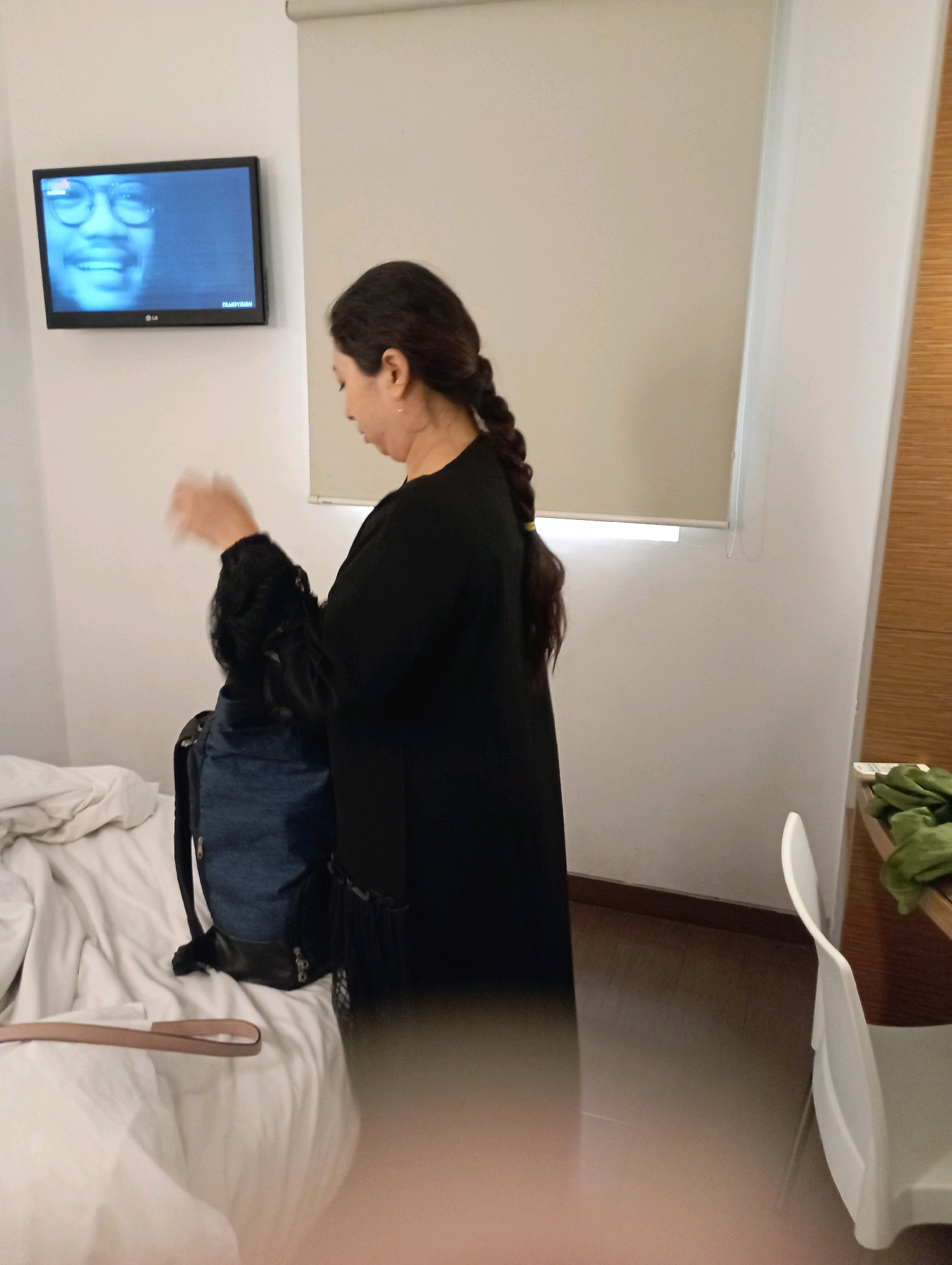 The image depicts an indoor scene, likely a hotel room in Malang, Indonesia. A woman with long, dark hair in a braid is seen from the side, wearing a black abaya. She appears to be adjusting something in her hands near a blue backpack. In the background, a flat-screen television mounted on a white wall displays a close-up of a smiling man wearing glasses. The room has white walls, a white bed with rumpled sheets, and a wooden-paneled wall to the right. A white chair and a desk with green fabric are also visible on the right side of the frame. The lighting suggests an indoor environment.
