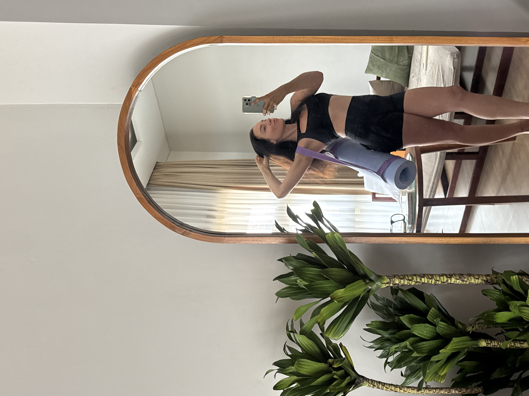A woman is standing and taking a mirror selfie in a room, likely a hotel room in North Kuta, Indonesia, judging by the architectural style. She is wearing a black sports bra and black athletic shorts, with a rolled-up purple yoga mat slung over her shoulder. Her right hand is holding a smartphone, and her left hand is touching her hair. The room has neutral-colored walls, with a large arched mirror on the right side. To the left of the mirror, there are three potted plants with long, green leaves. In the reflection of the mirror, a window with sheer curtains, a wooden table, and a sofa with pillows can be seen.