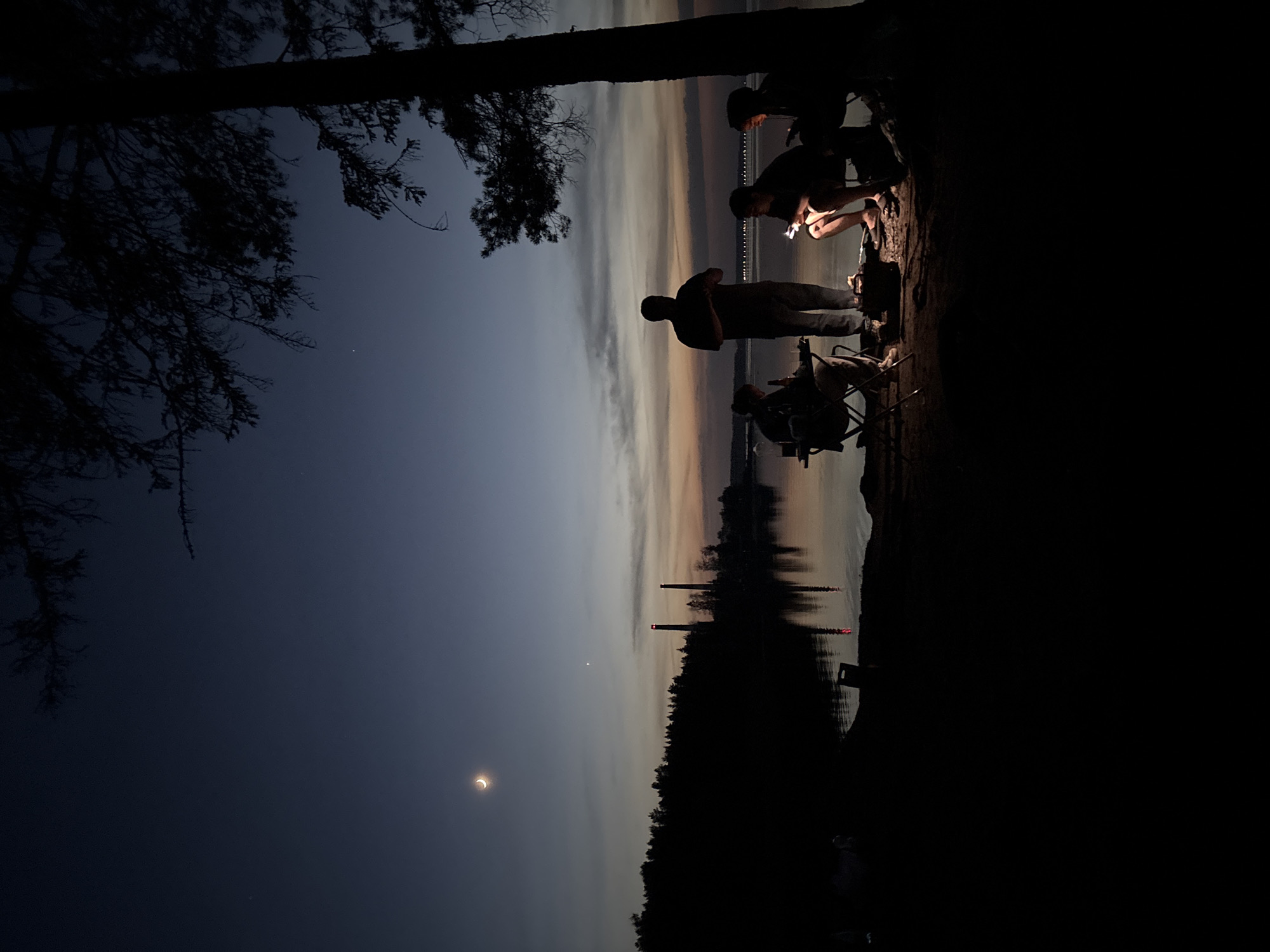 The image, taken in a low-light setting possibly at dusk or dawn, depicts a group of people silhouetted against a body of water. The location is Chaikovsky, Russia, as indicated by the presence of what appears to be the "FOSTEK" boiler stacks. The people are positioned on an elevated shoreline. A crescent moon is visible in the sky on the left. In the background there are lights on the water and trees. A tree is in the far left corner.