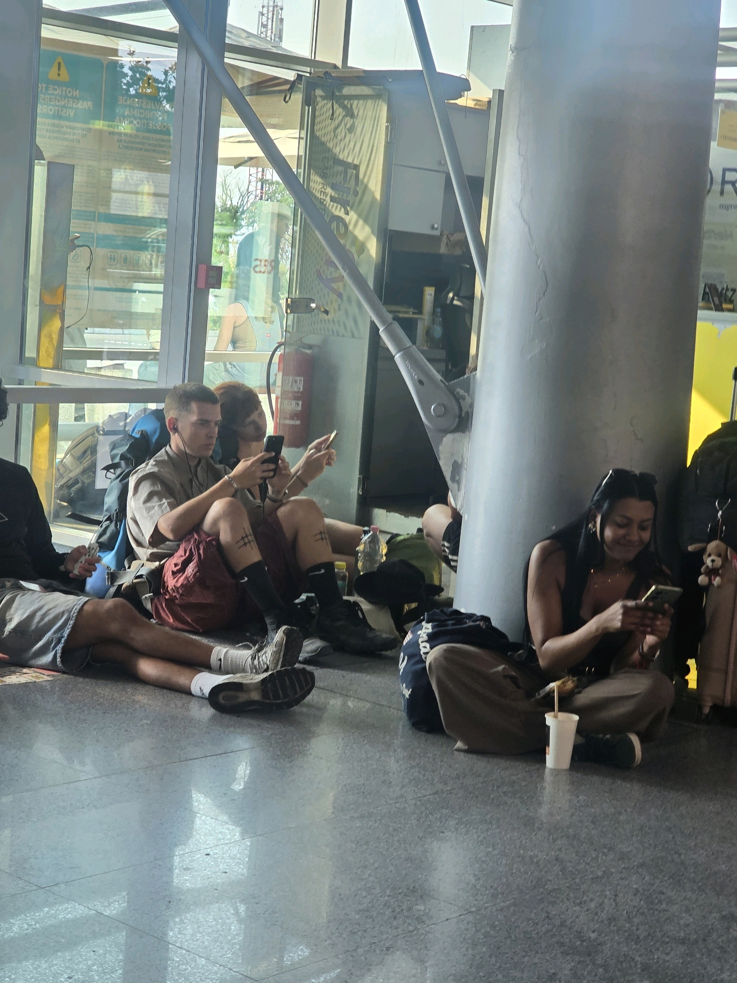 In Golubovci, Montenegro, at what appears to be an airport terminal, a group of individuals are seated on the floor, resting their luggage around them. The floor is made of polished gray tiles, reflecting the light from large windows and overhead fixtures.
In the foreground, a young woman with long dark hair is engrossed in her smartphone, her legs crossed and wearing khaki-colored pants and black sneakers. Beside her, a white cup with a straw rests on the floor. To her left, a young man in a light brown shirt and dark red shorts is also looking at his phone, wearing black socks and sneakers. He has a blue backpack behind him and appears to be sitting with another person whose back is to the camera, wearing a light blue tank top and grey shorts. Further to the left, another person's legs are visible, wearing grey shorts and white socks.
The background shows large glass windows that allow natural light to enter, revealing trees and a building structure outside. A fire extinguisher is visible mounted on a wall. The overall atmosphere suggests a period of waiting or transit within the terminal.